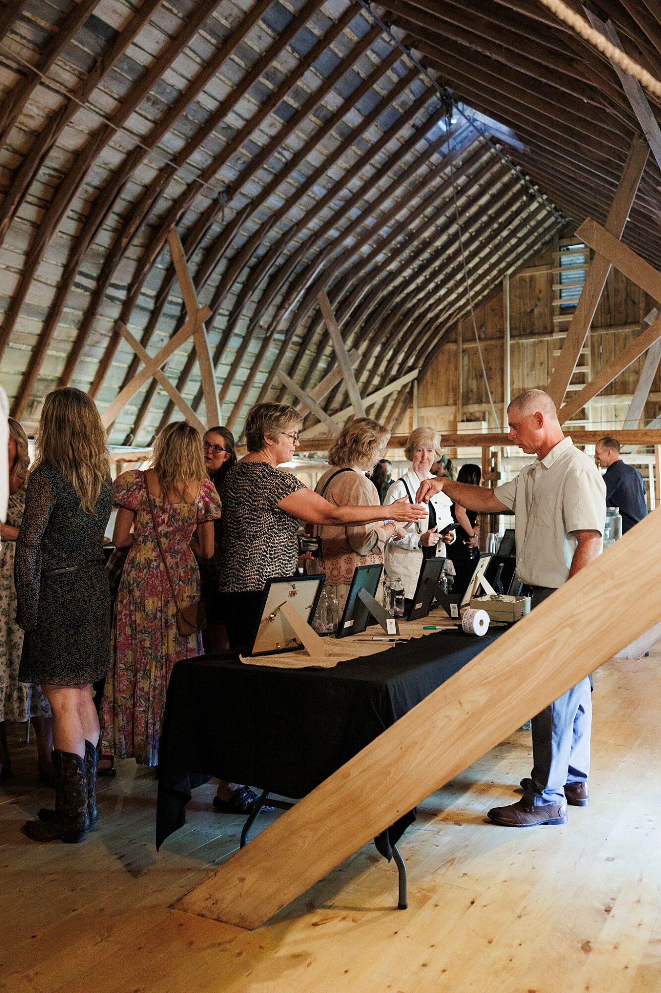 Group of people gathered around a table inside a wooden structure, with a man handing out items. Wooden beams and floor visible.
