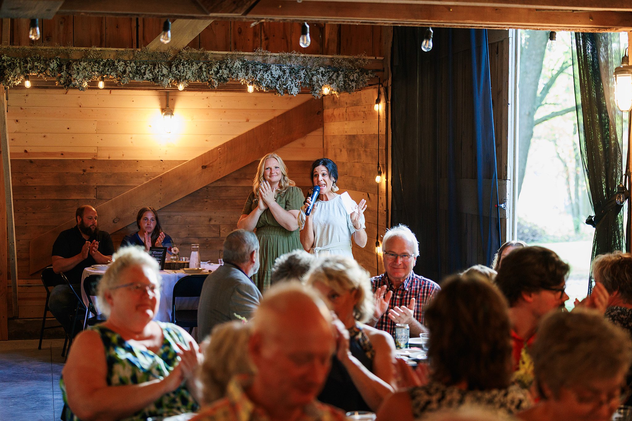 People gathered around tables in a rustic indoor setting with wooden walls and large windows, some standing and singing.