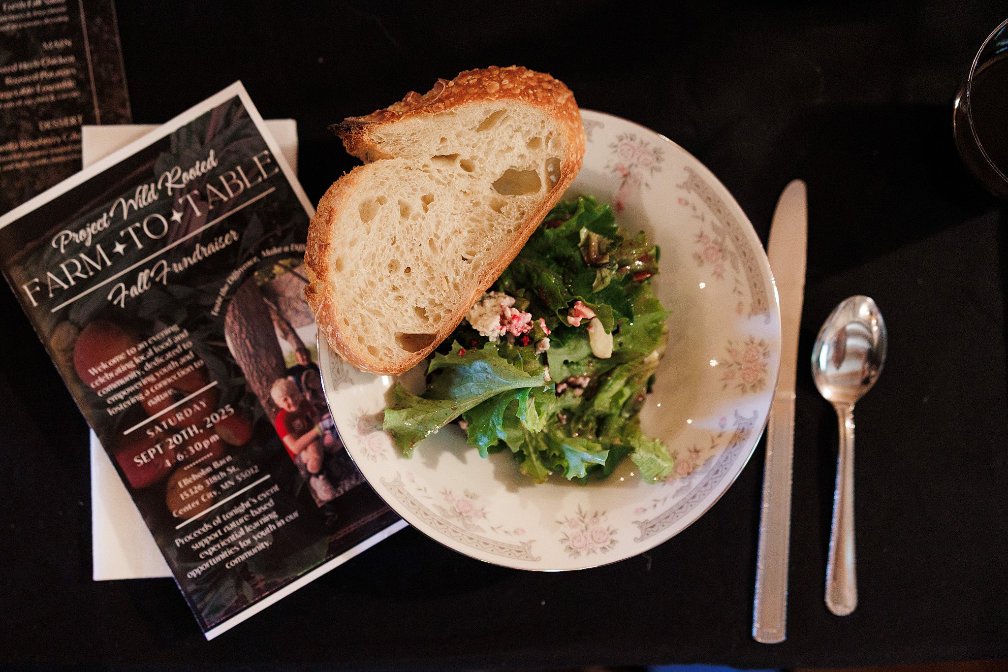 A plate with a slice of bread and a small salad with greens and flowers, a menu, a knife, and a spoon on a black table.