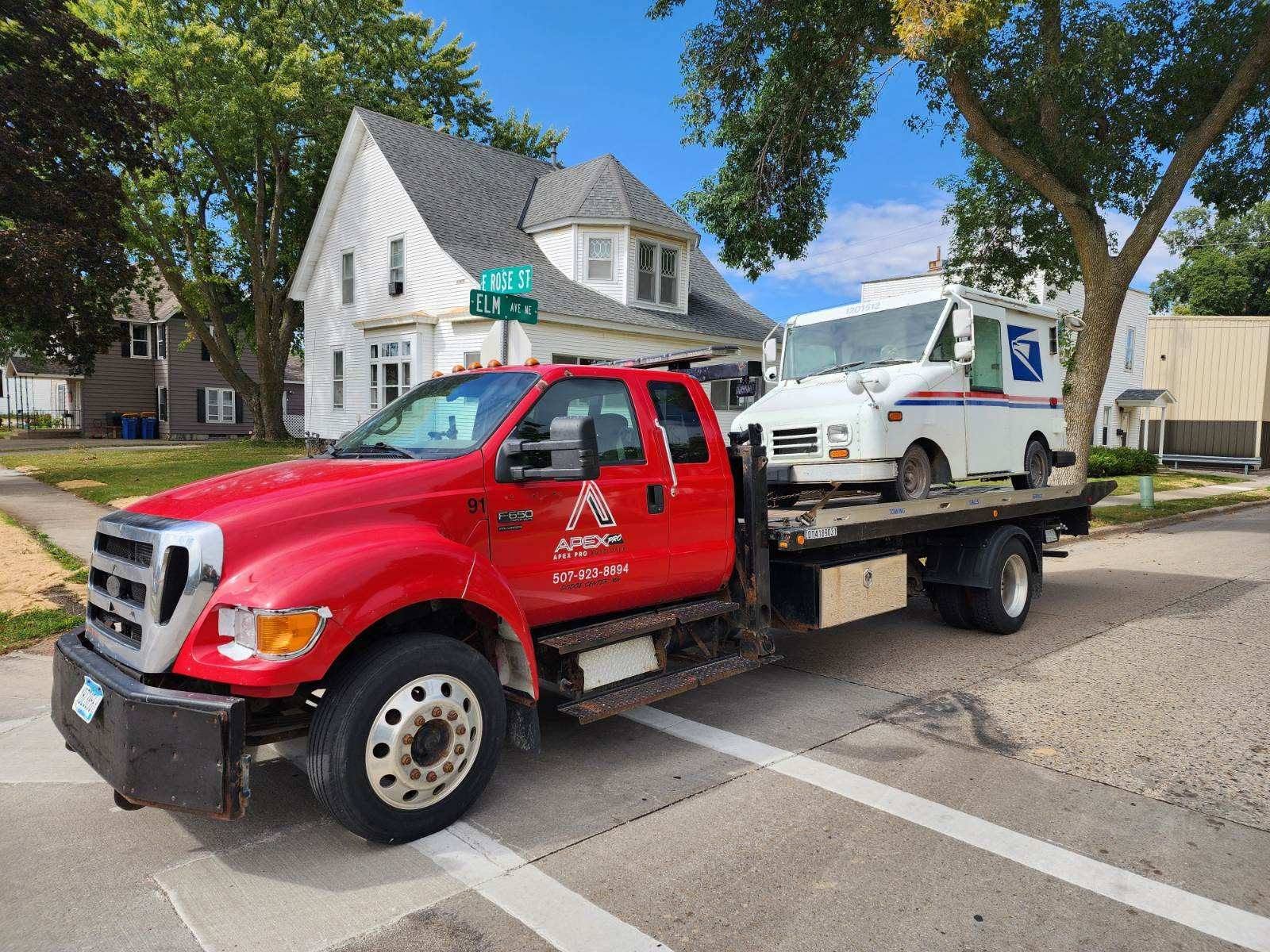 Red tow truck with USPS vehicle on flatbed, parked on street with houses and trees in background.