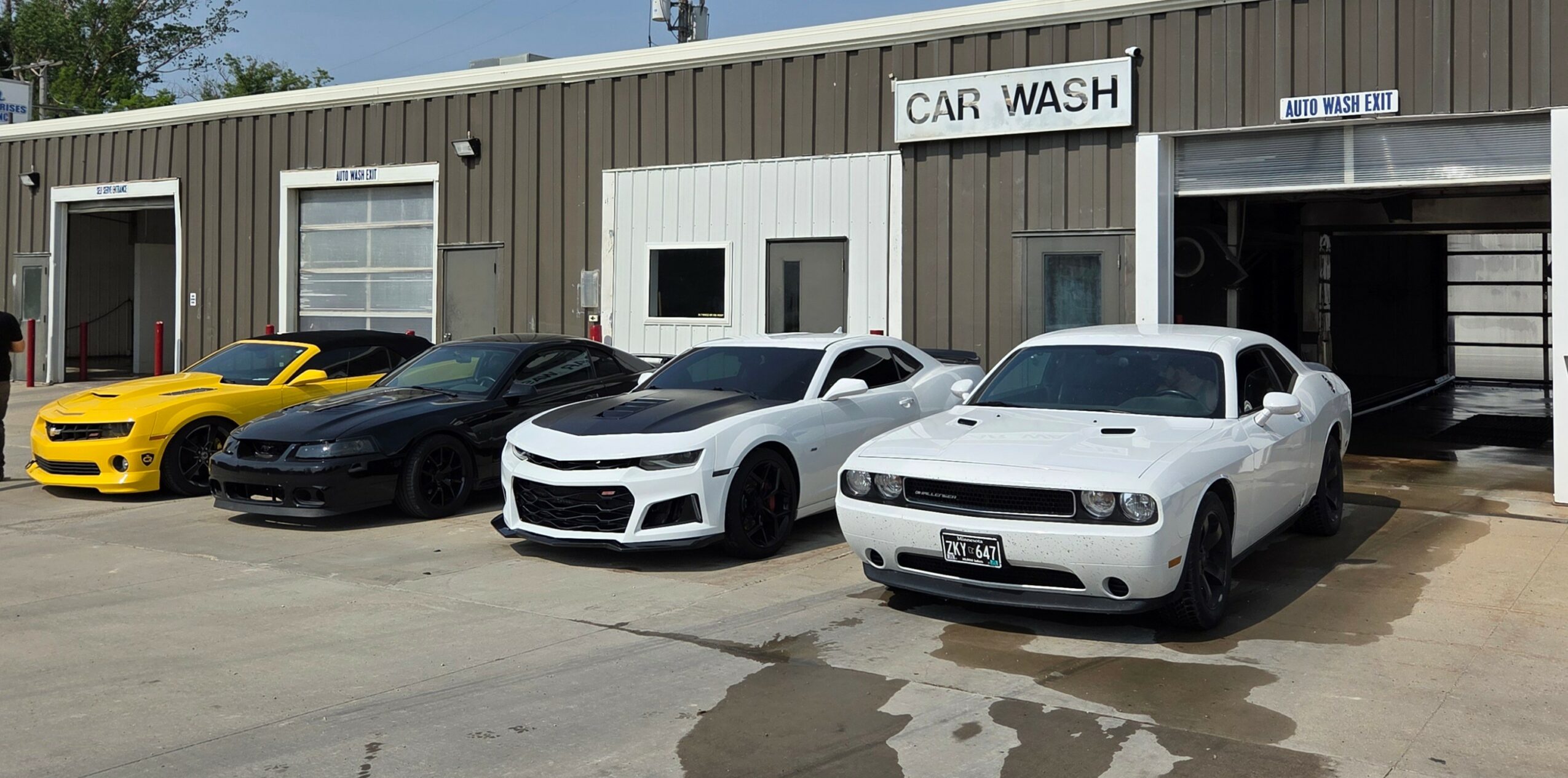 Four cars parked outside a car wash building, including a yellow sports car, black car, white car, and gray car.