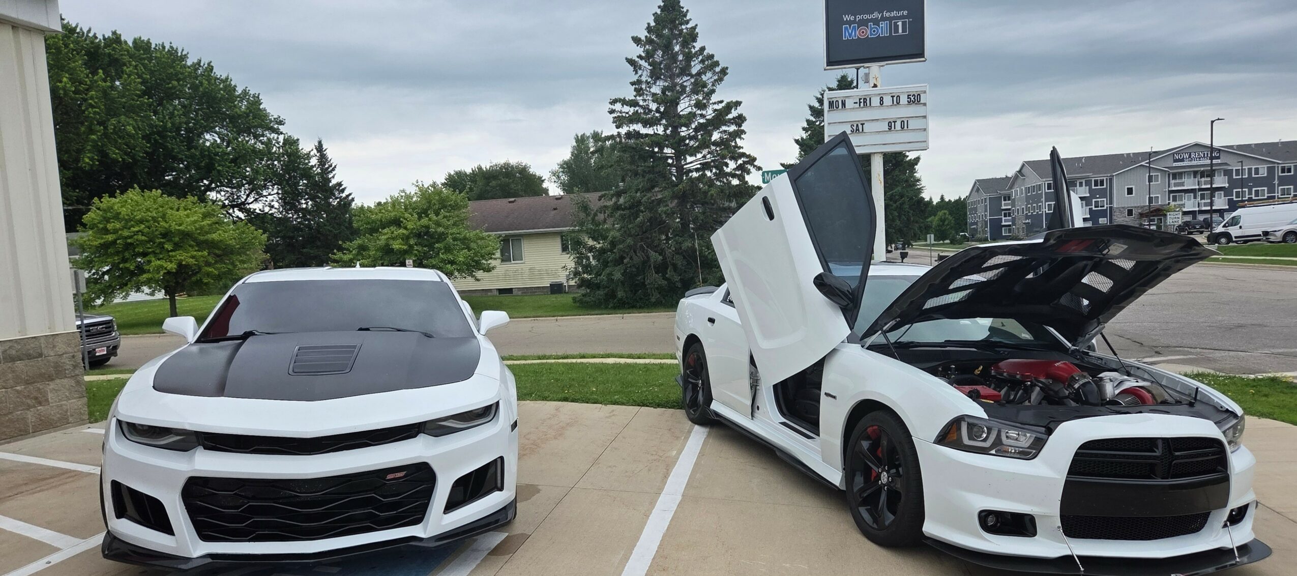 Two white sports cars parked side by side in a parking lot, one with its doors open, engine visible.
