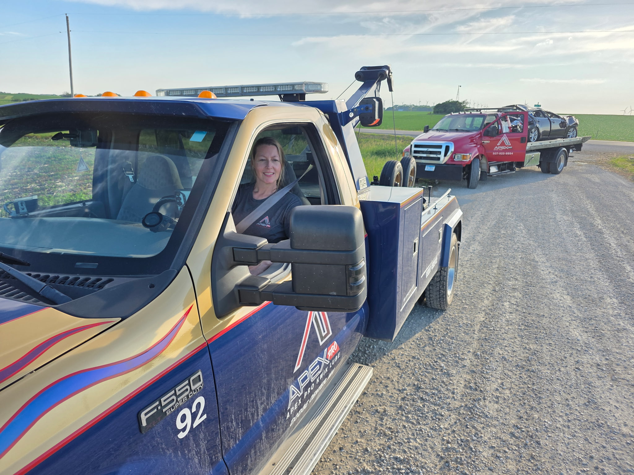 Police pickup truck with a person inside, followed by a red pickup truck towing a black car on a rural road under a blue sky.