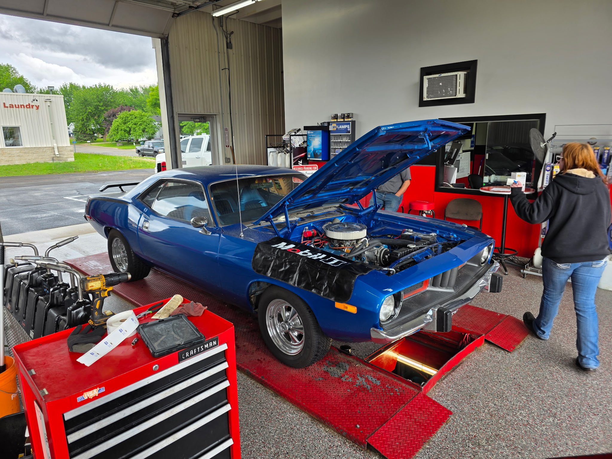 Blue classic car with open hood inside a garage, person working on it, tools and equipment nearby.