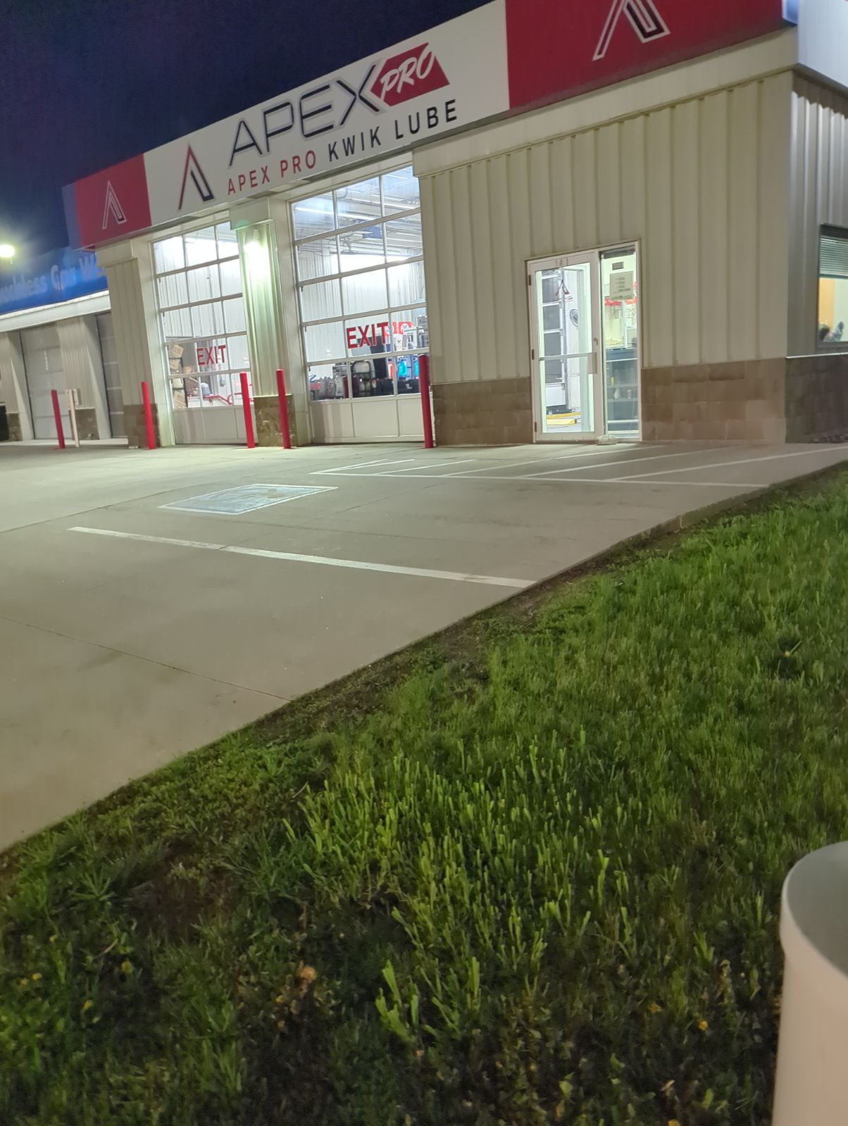 Storefront with large windows and a door, parking lot in front, and a grassy area to the right, evening lighting.