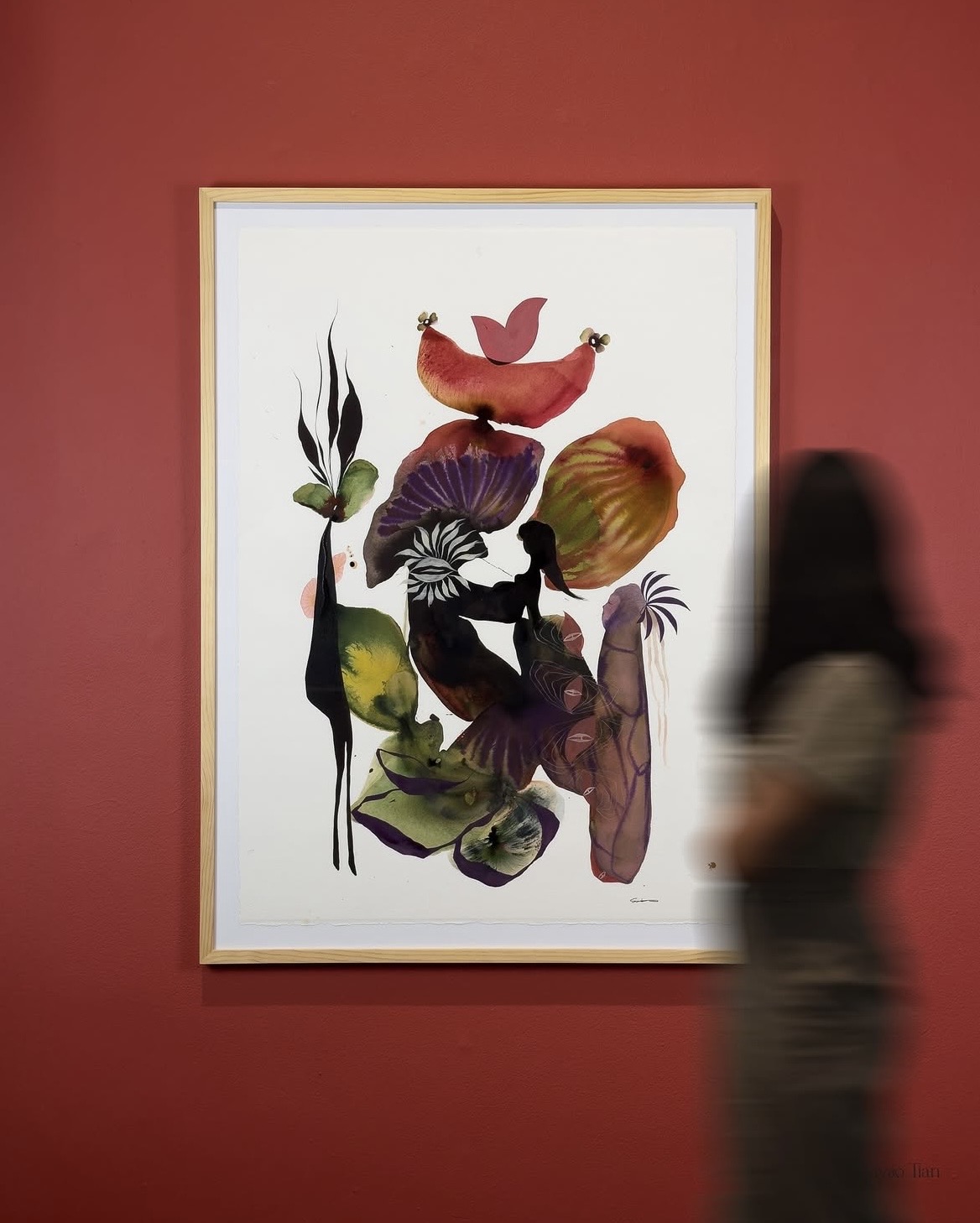 Colorful abstract artwork with various shapes and plant-like elements on a white background, framed and hung on a red wall.