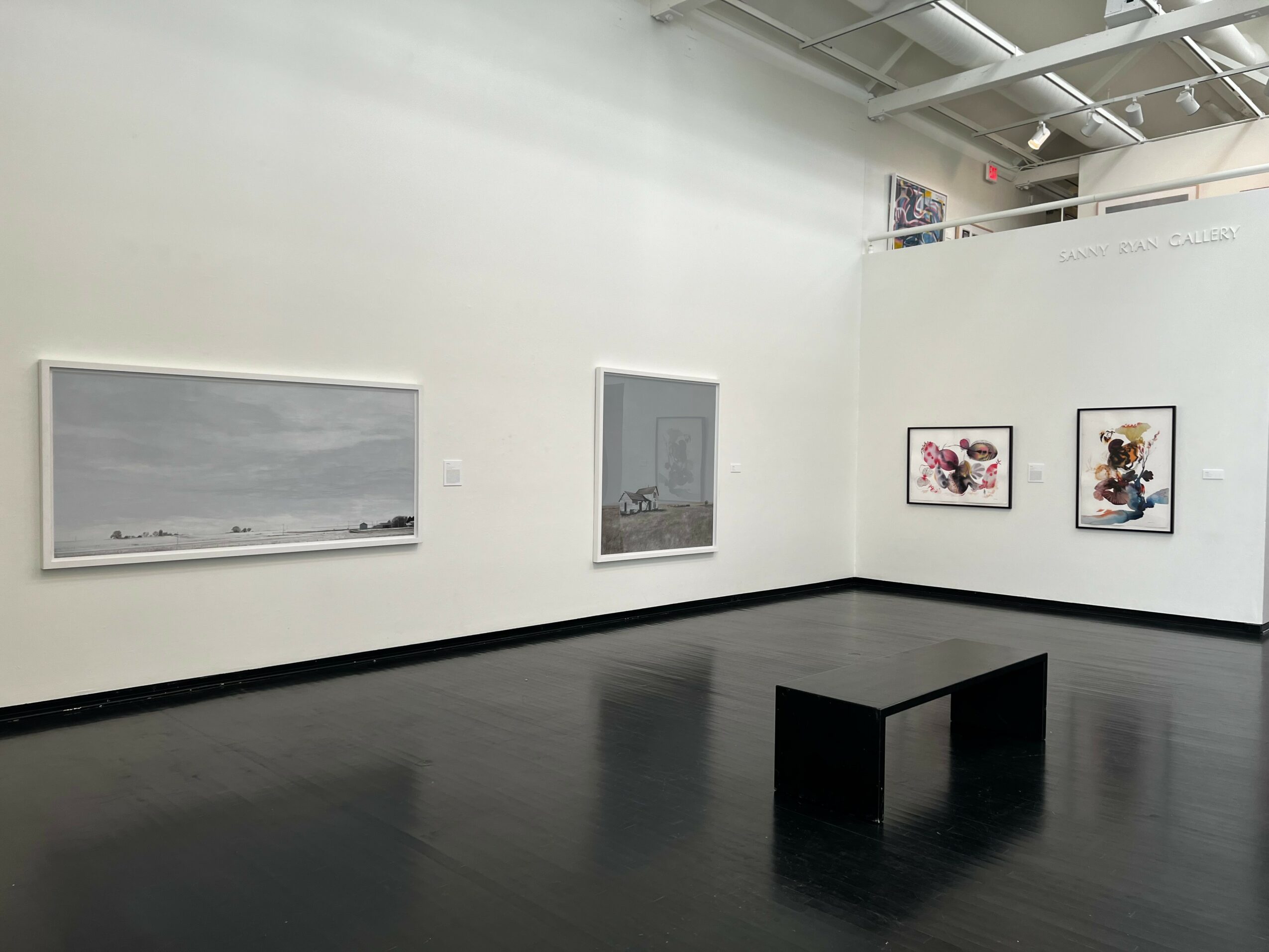 Gallery with framed artworks on white walls and a black bench in the center, ceiling with exposed beams.