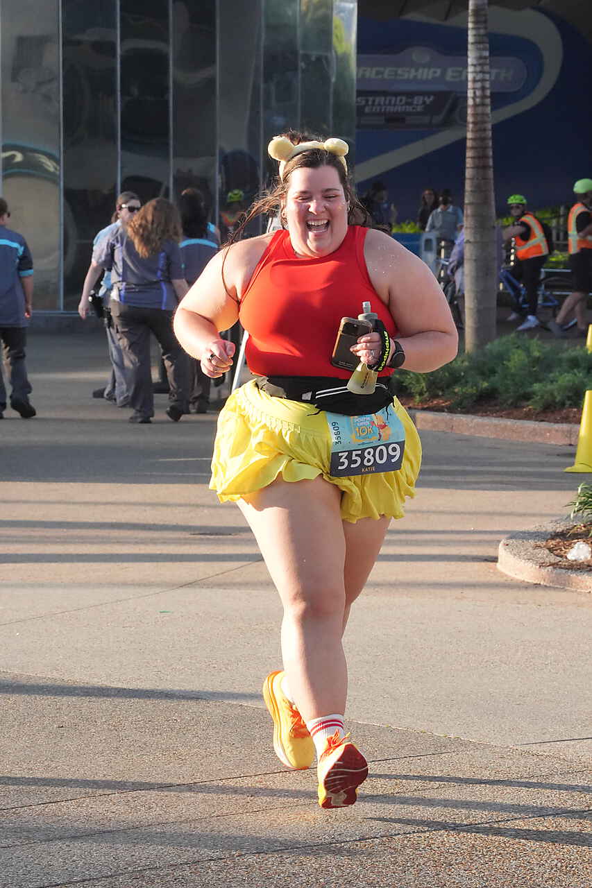 Smiling woman running in a race, wearing a red top, yellow tutu, and striped socks, holding a phone, with spectators in background.