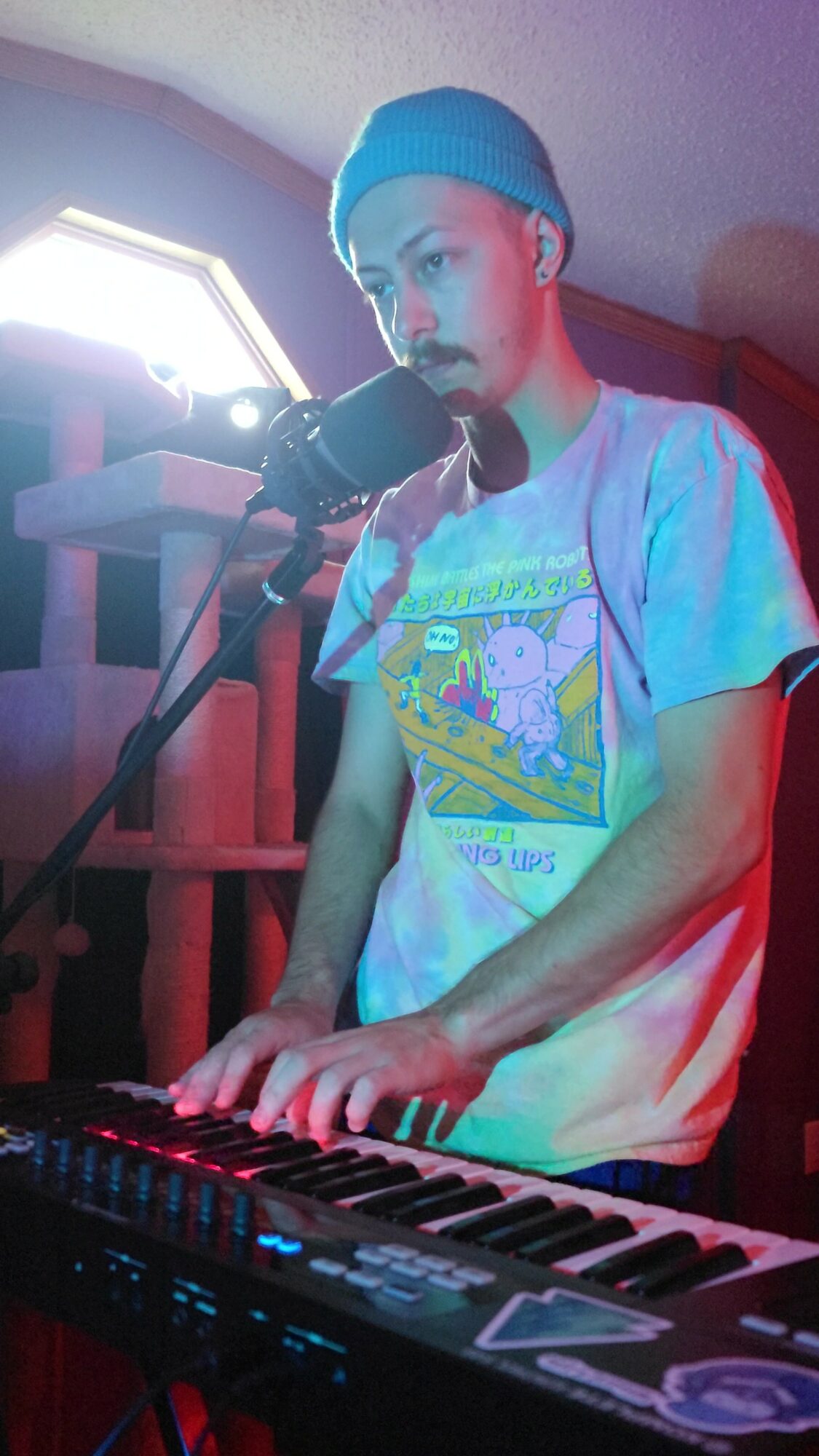 Young person wearing a blue beanie playing keyboard with microphone, colorful lighting, and a window in the background.
