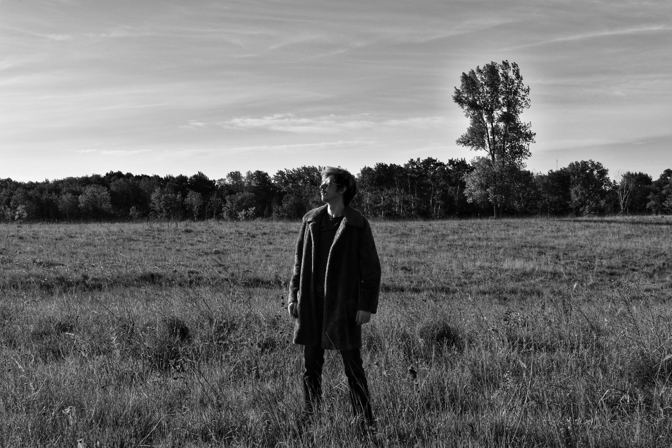Person standing in a field with trees and sky in the background, black and white image.