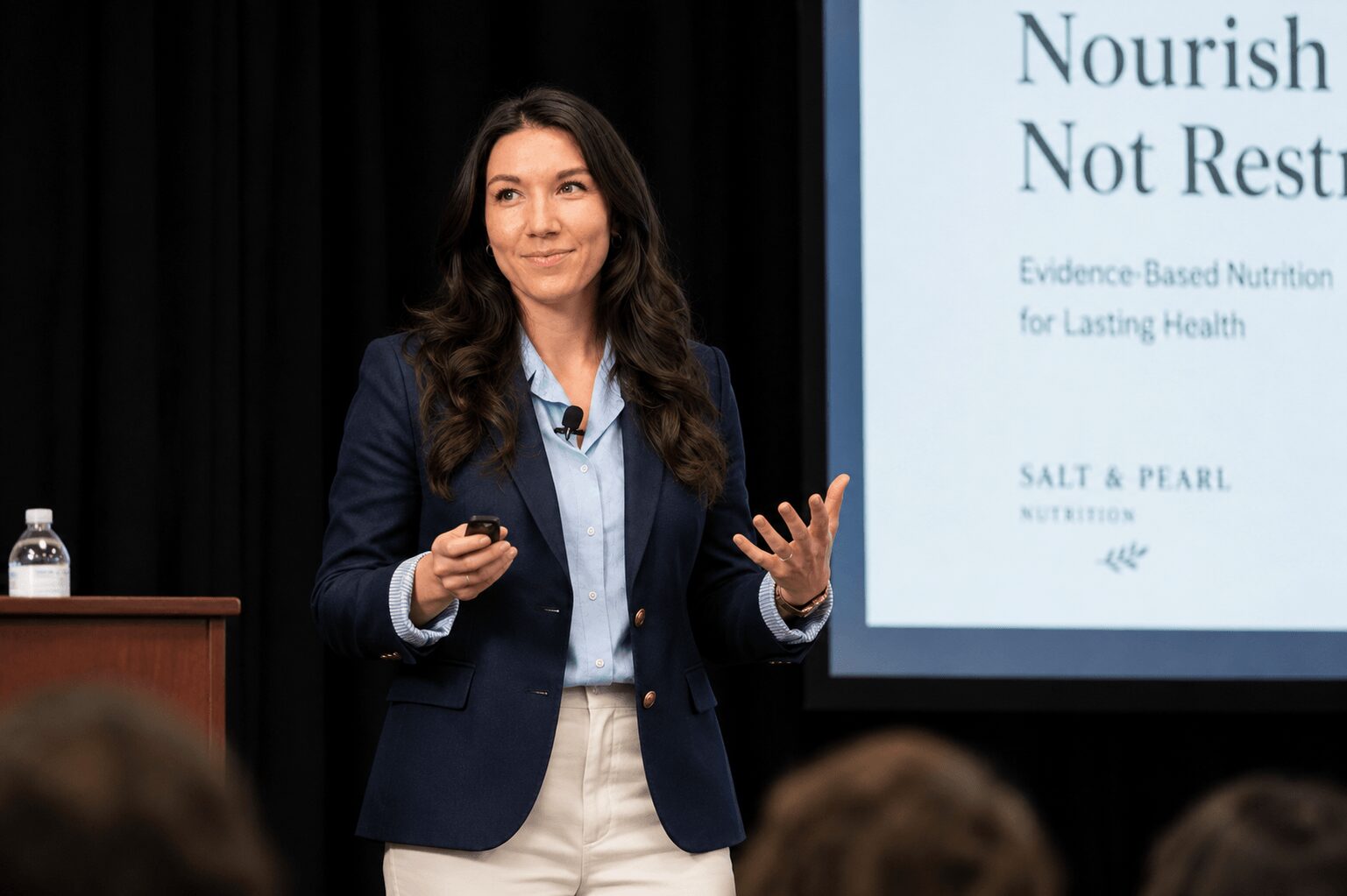 Woman in a dark blazer and light shirt speaking in front of a presentation screen, audience in foreground.