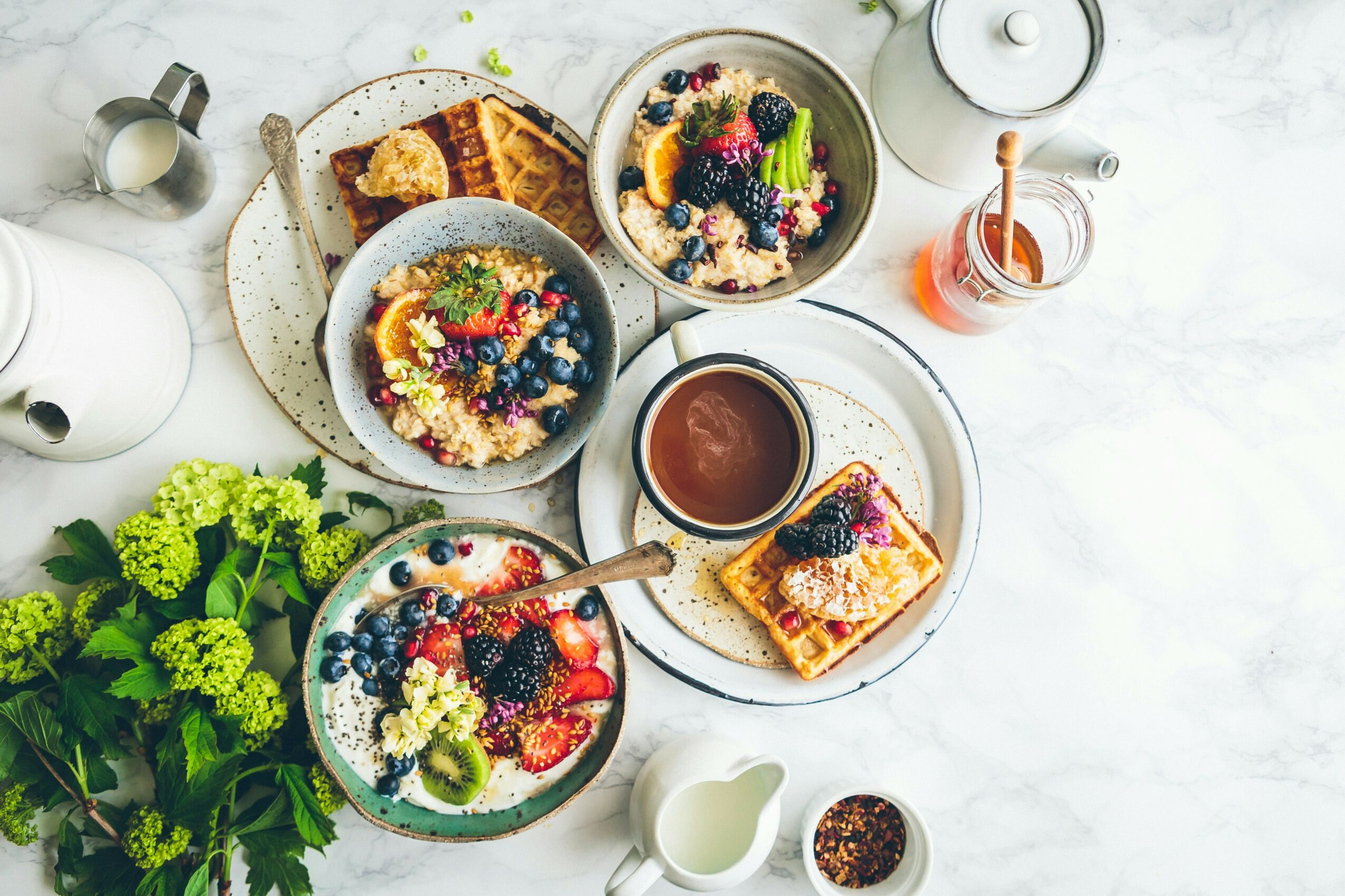 Breakfast spread with bowls of fruit, granola, toast, a cup of tea, and a small jar of jam on a white surface.