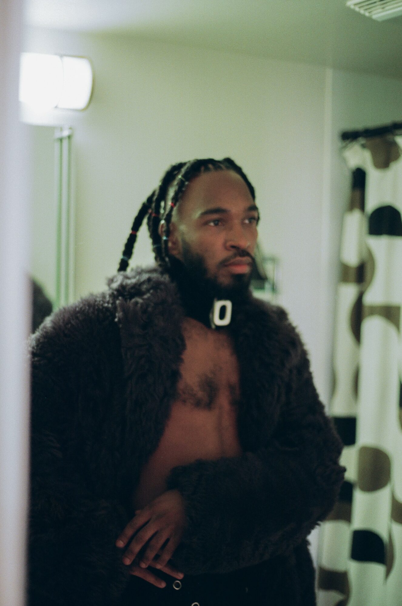 Man with braided hair and beard, shirtless, wearing a dark fur coat, standing indoors near a window and curtain.