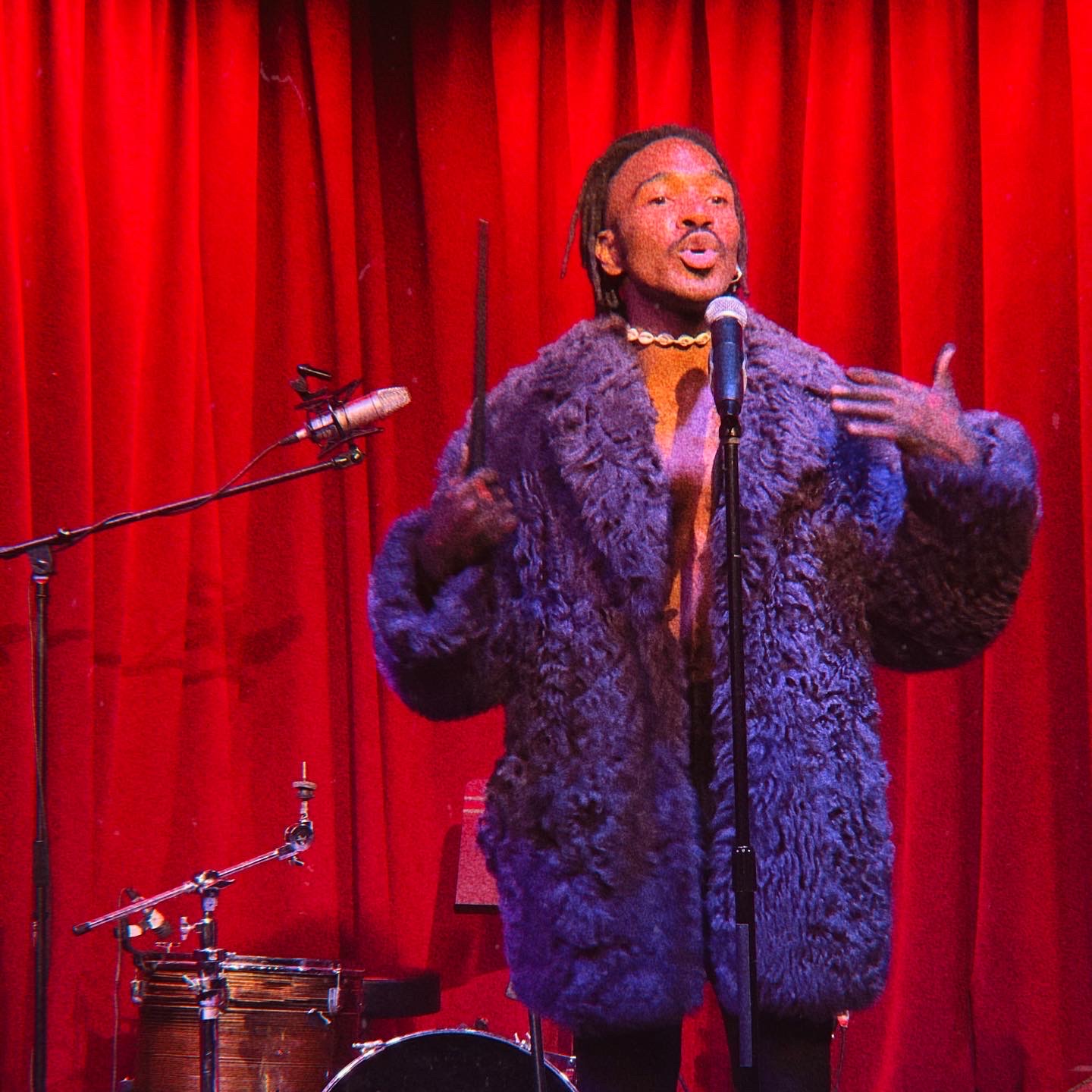 Person performing on stage with red curtain background, wearing a large fur coat and speaking into a microphone.