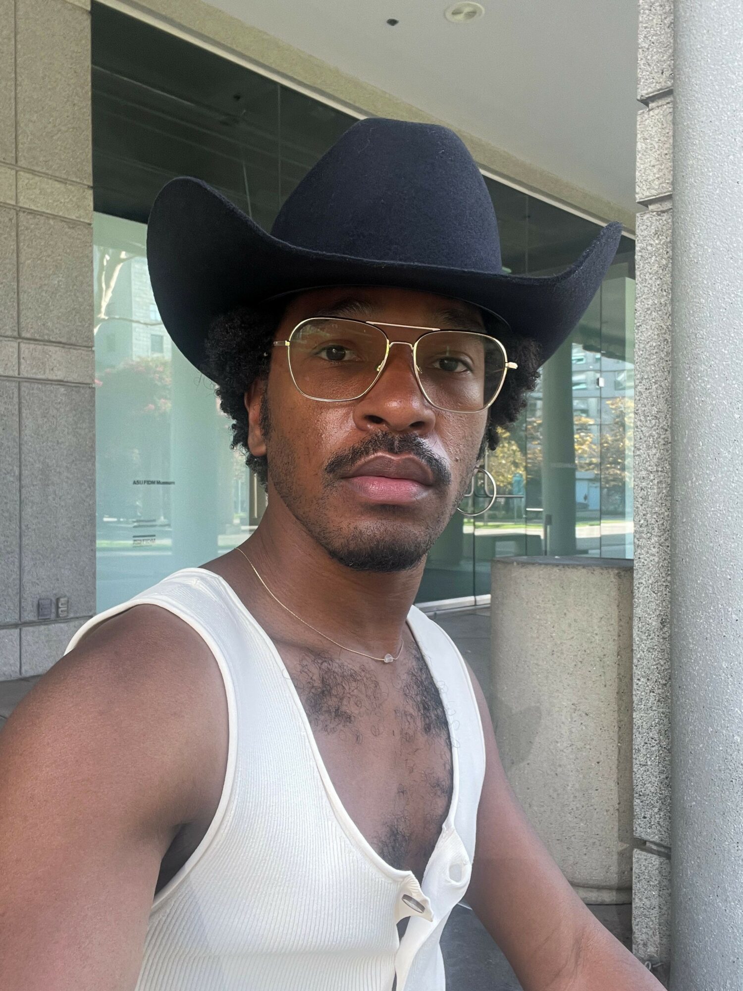 Person wearing a black hat, glasses, and a white sleeveless top, standing outdoors with a cityscape background.