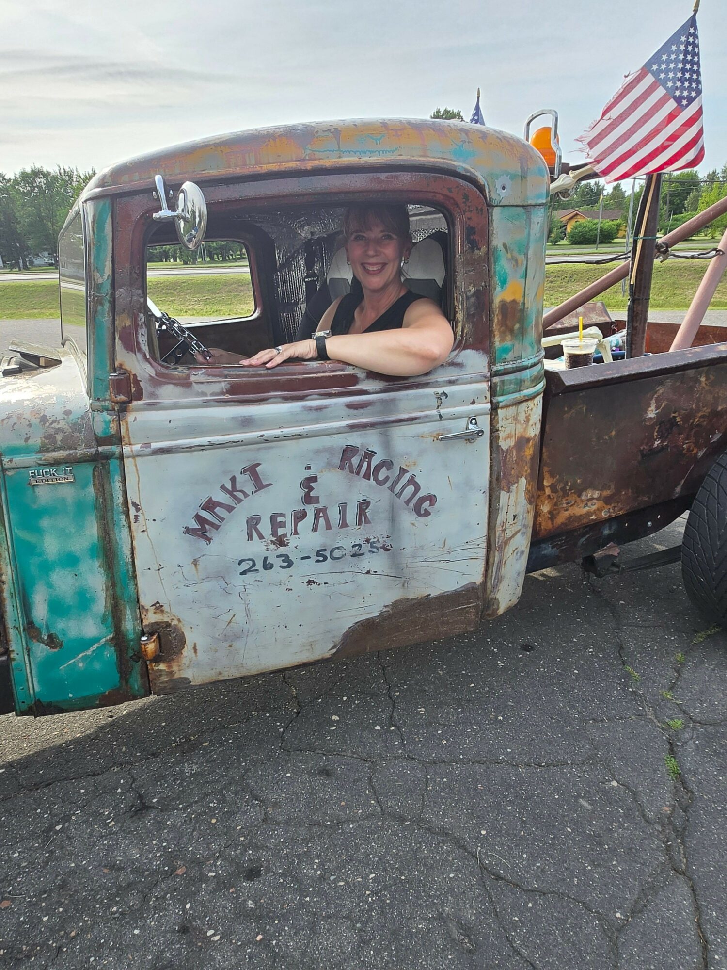 Person sitting inside an old, rusty truck with a sign reading 'The Olde Reptile' on the door, outdoors with American flags in background.