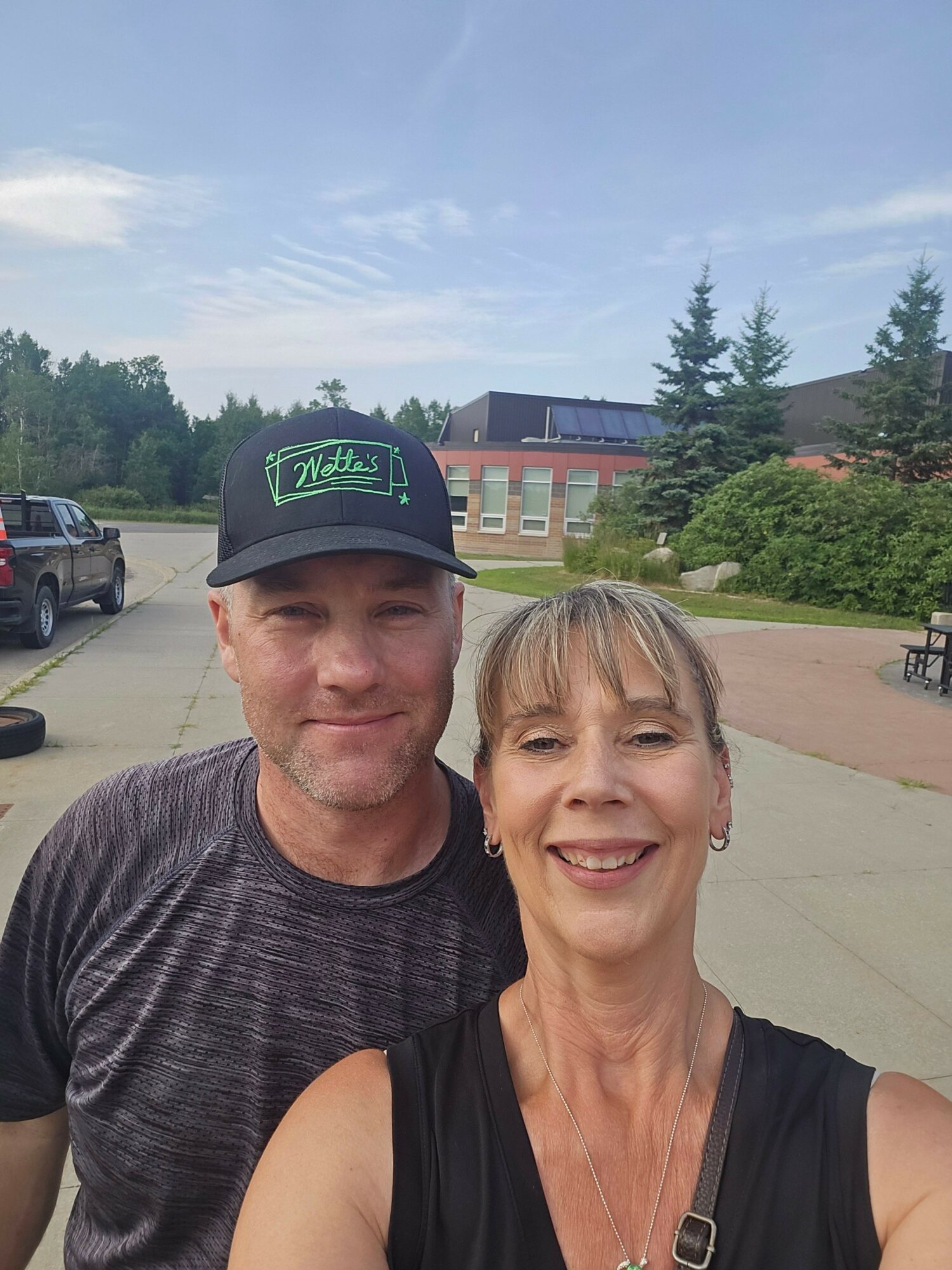 Two people standing outdoors, one wearing a cap and the other smiling, with trees and buildings in the background.