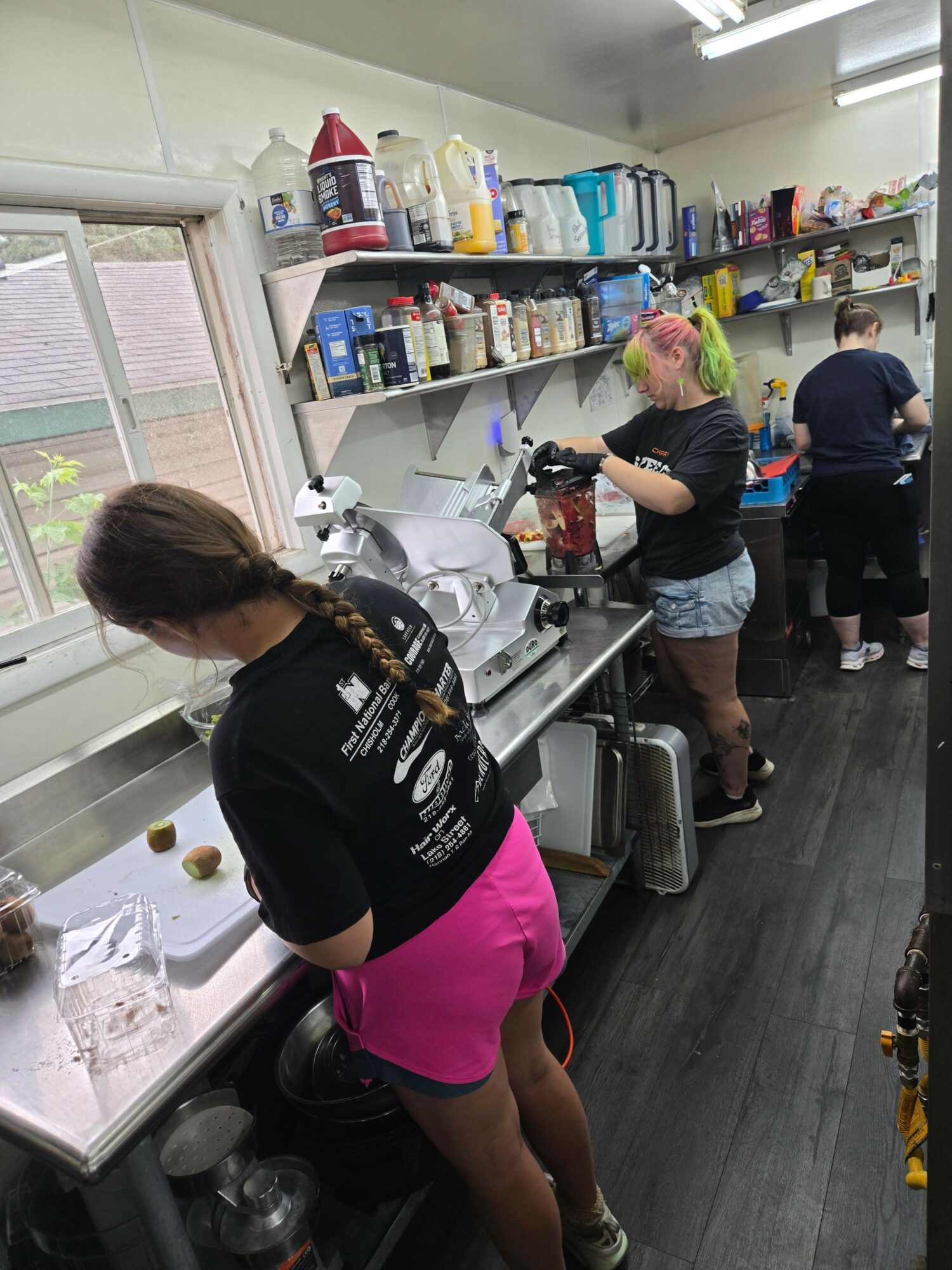 People working behind a counter in a busy kitchen or cafe, with shelves of supplies and equipment visible.