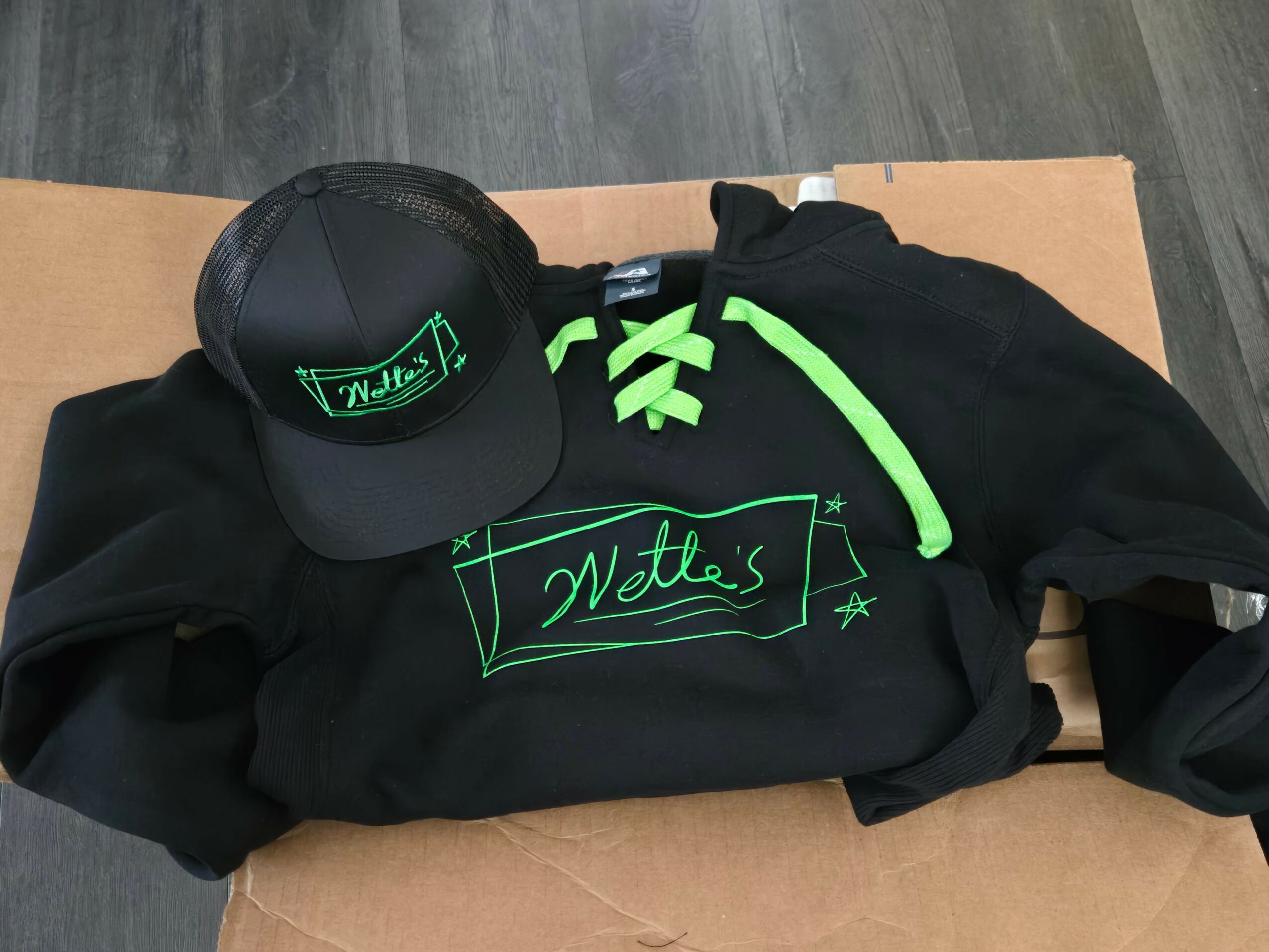 Black hoodie with green drawstrings and green writing, a black cap with green design, laid on a cardboard surface.