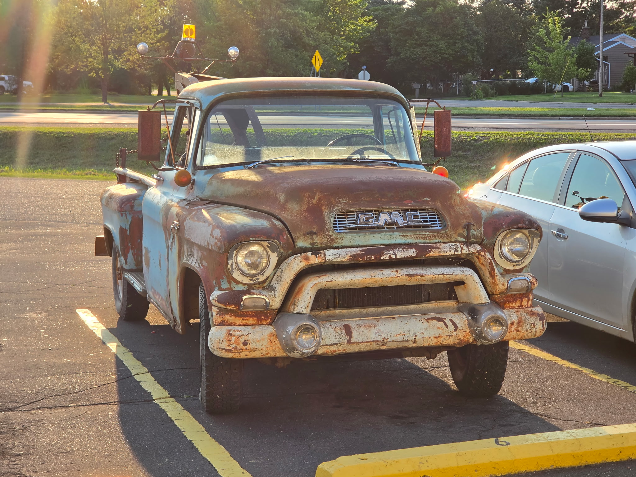 Rusty, old pickup truck parked in a lot with a silver car beside it, trees and street in background.