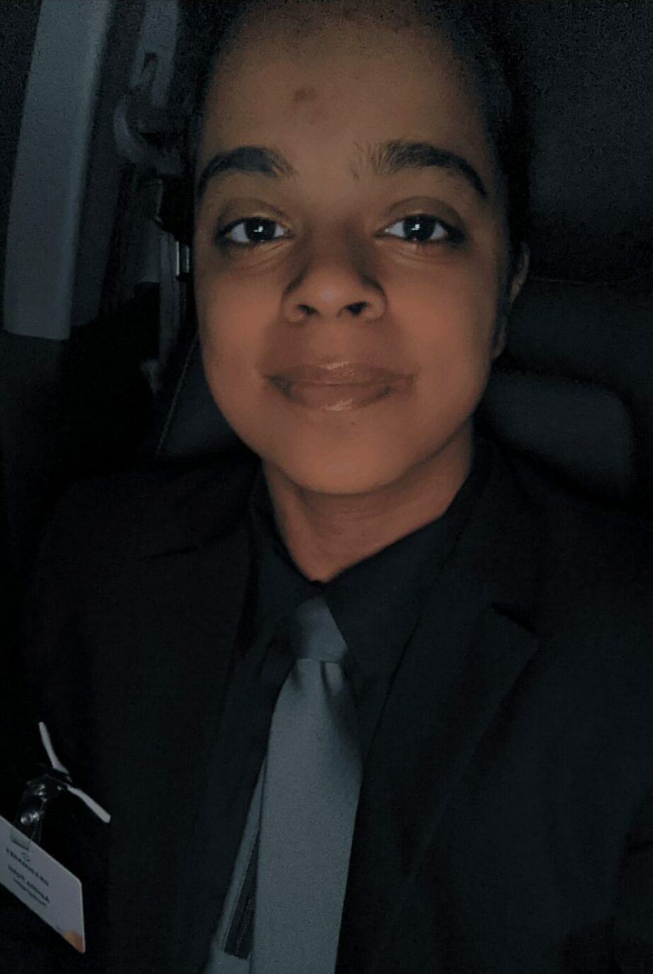 Person with dark hair and dark eyes wearing a black suit and gray tie, smiling slightly, in a dimly lit environment.