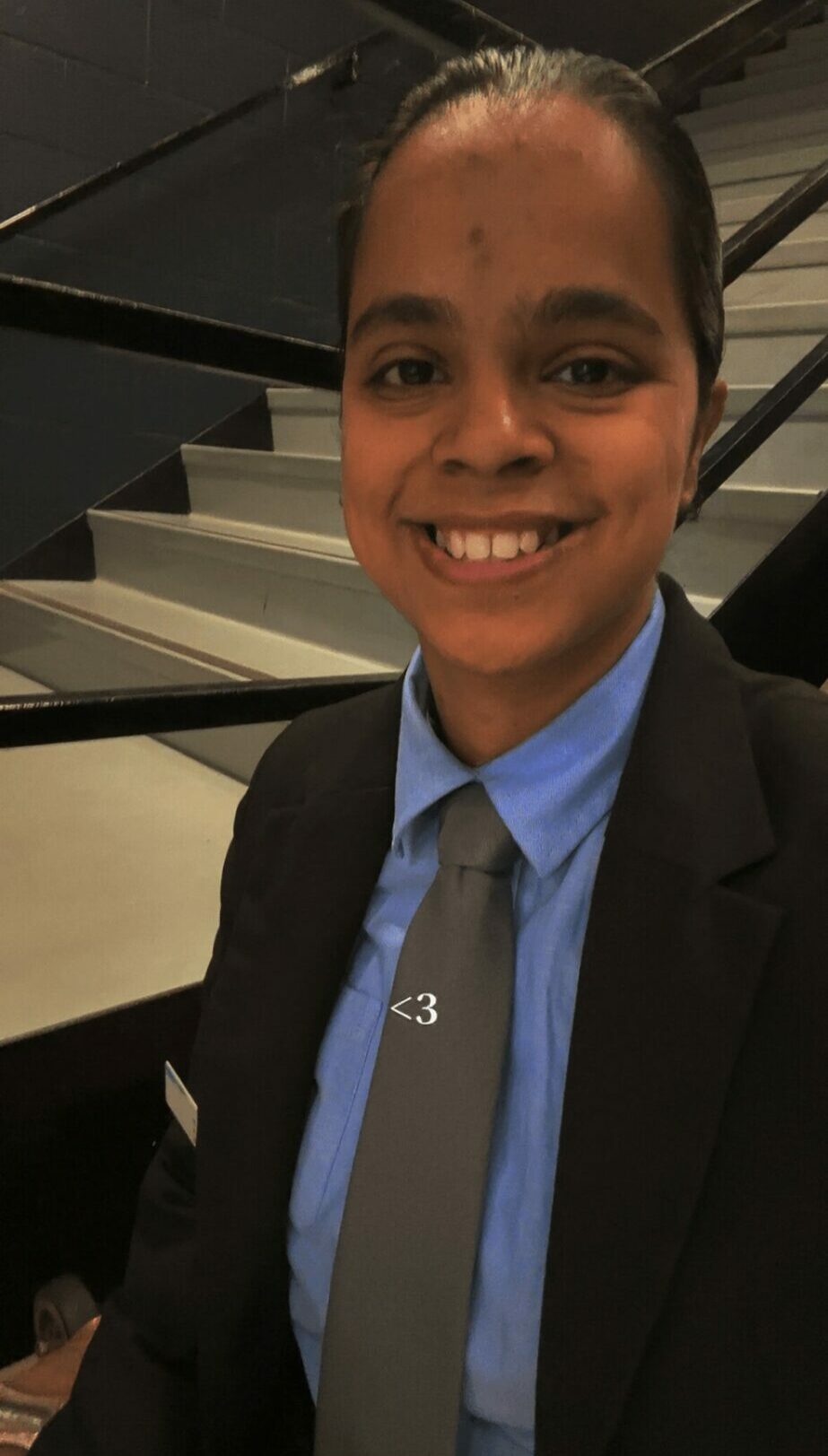 Young person smiling, wearing a suit and tie, standing indoors near stairs.