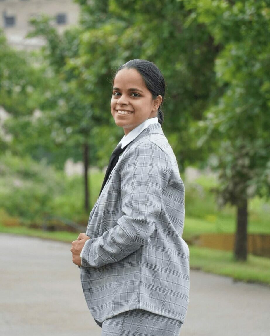 Young woman in gray blazer smiling outdoors with trees in background.