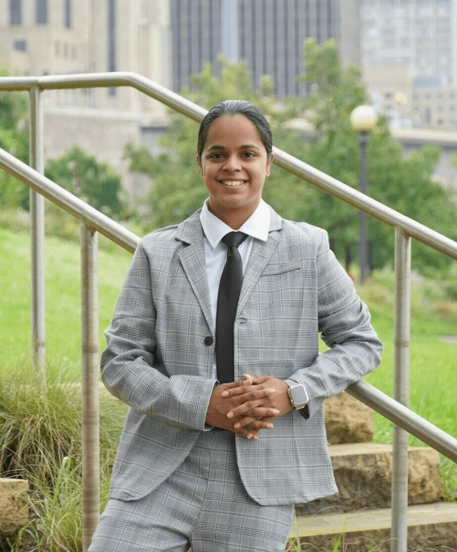 Woman in gray suit and tie standing outdoors with buildings and greenery in background.
