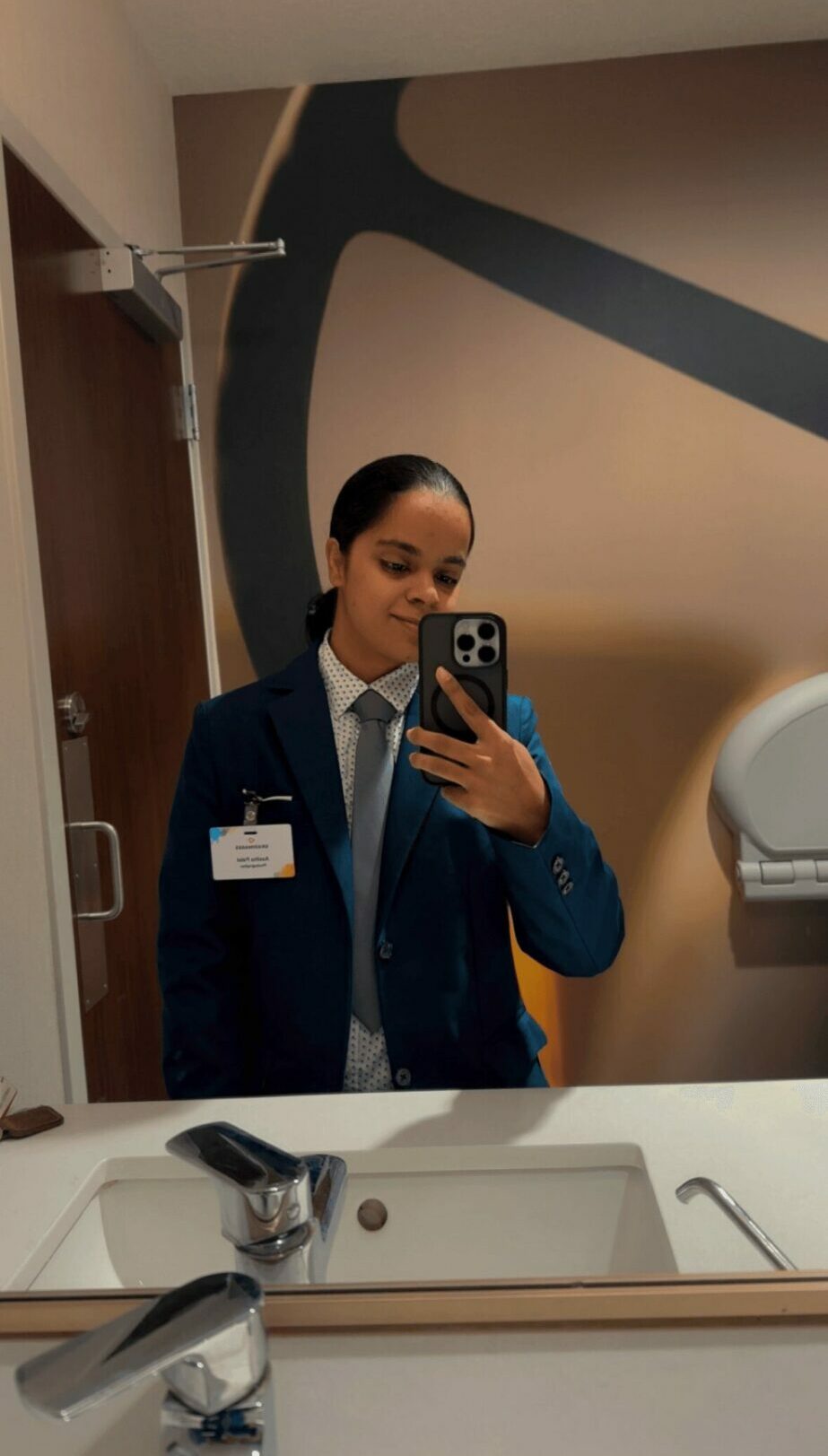 Person in a suit taking a mirror selfie in a bathroom, holding a phone, with a large abstract wall design behind.