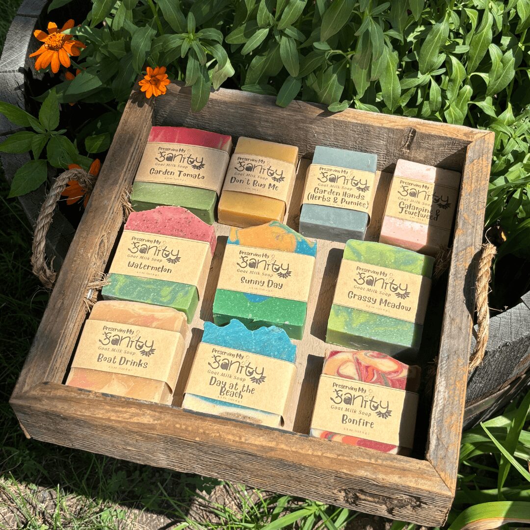 Wooden tray with colorful soap bars labeled with garden-themed names, placed outdoors among green plants and orange flowers.