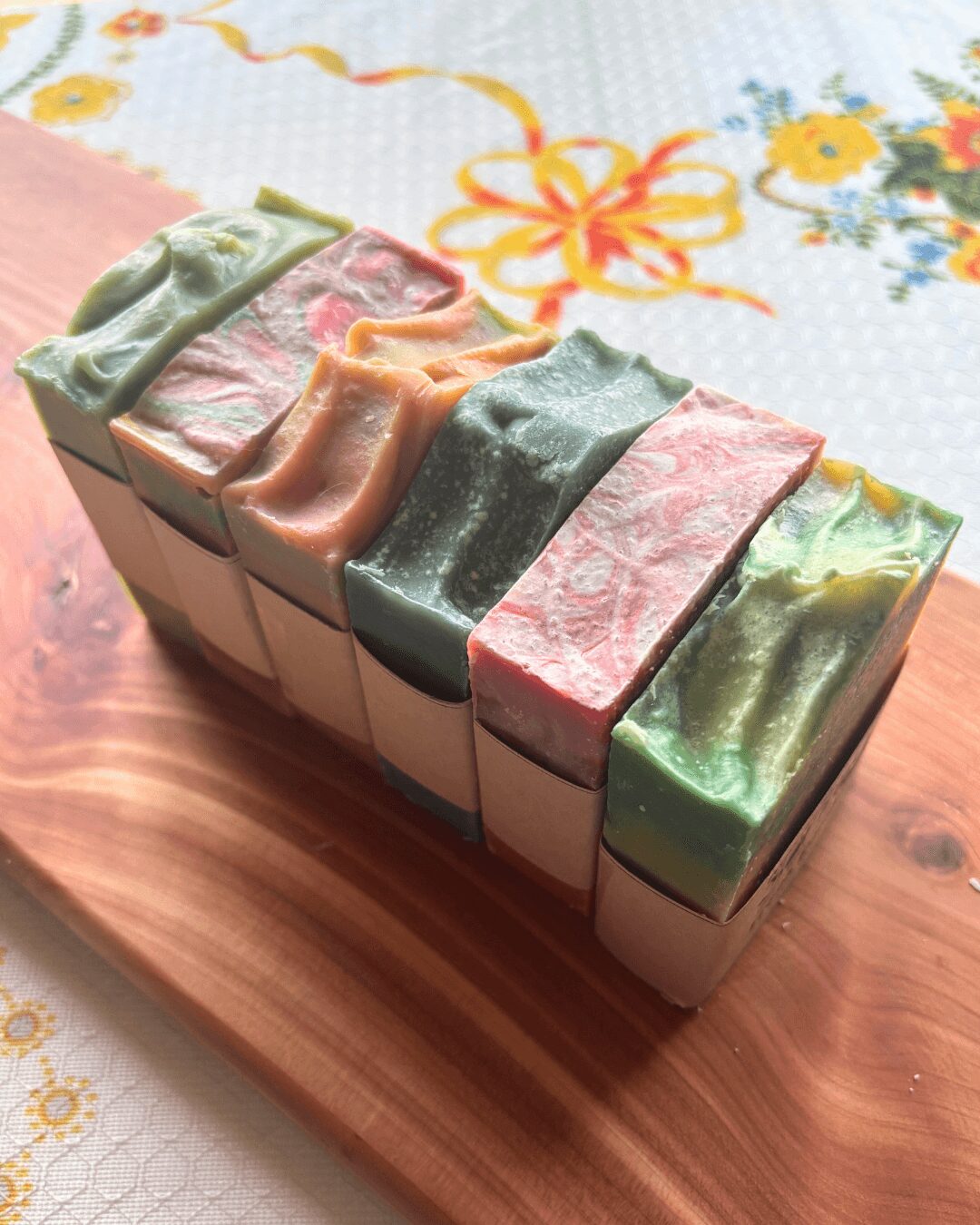 Six soap bars with colorful, marbled tops arranged on a wooden surface, with a floral tablecloth in the background.