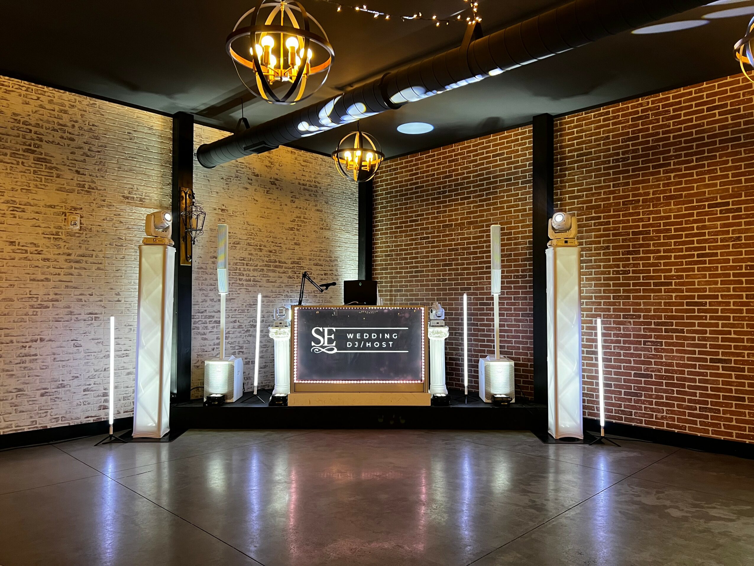 DJ booth with speakers and lighting setup against brick wall, in a dimly lit room with decorative ceiling lights.