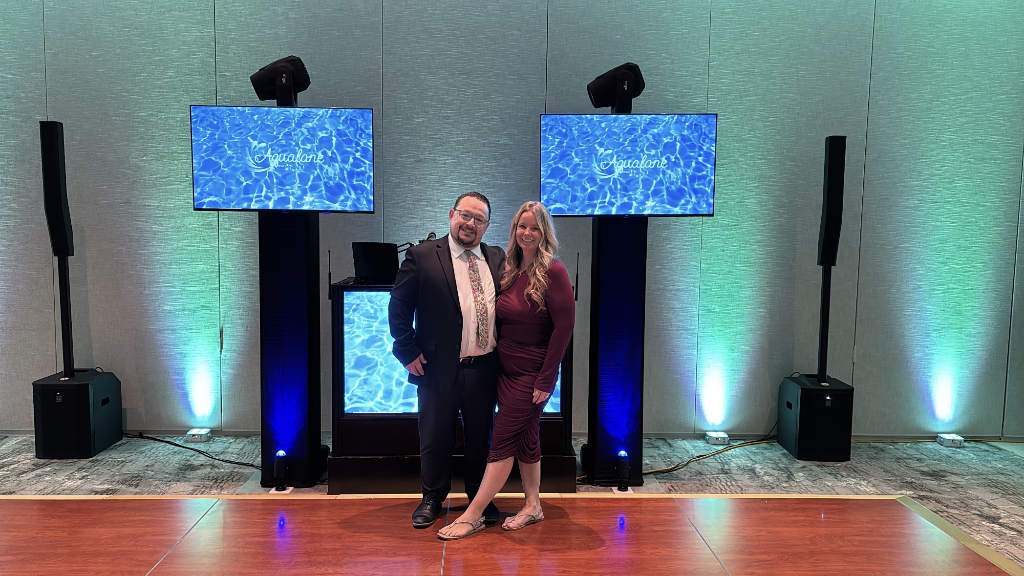 Two people stand in front of a stage with two large screens and speakers, smiling at the camera.
