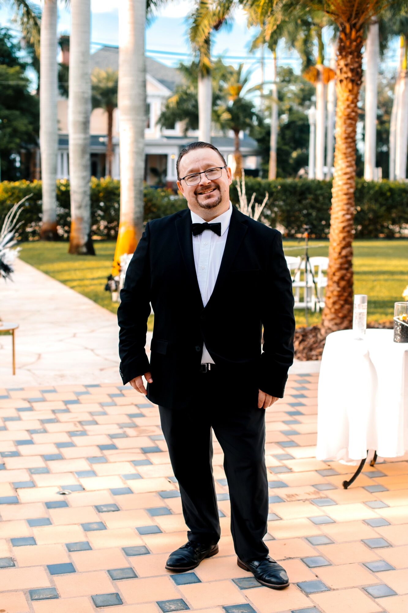 Man in tuxedo smiling outdoors with palm trees and buildings in background.