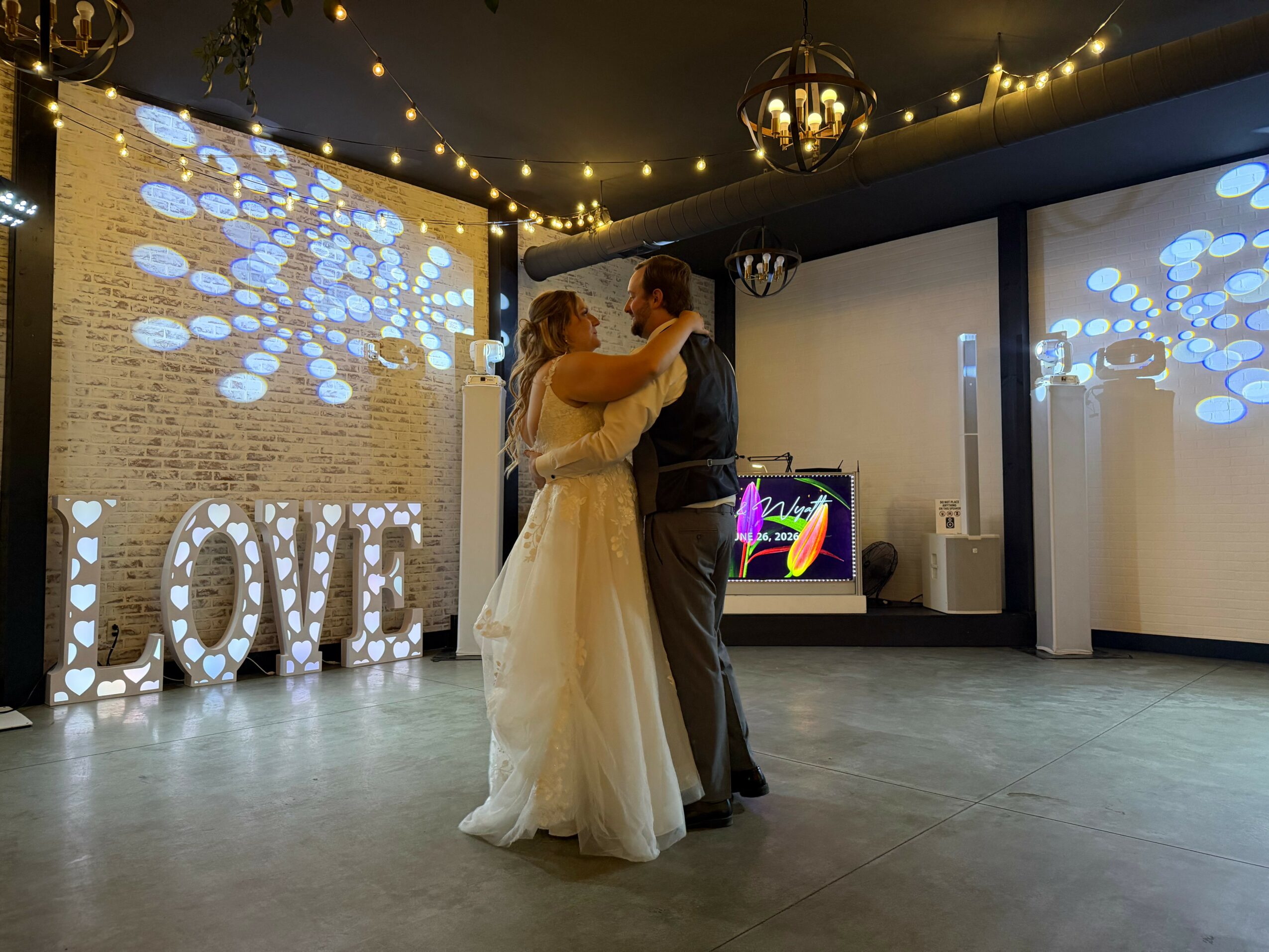 Couple dancing in a decorated indoor venue with string lights and illuminated decorations, including a large 'LOVE' sign.