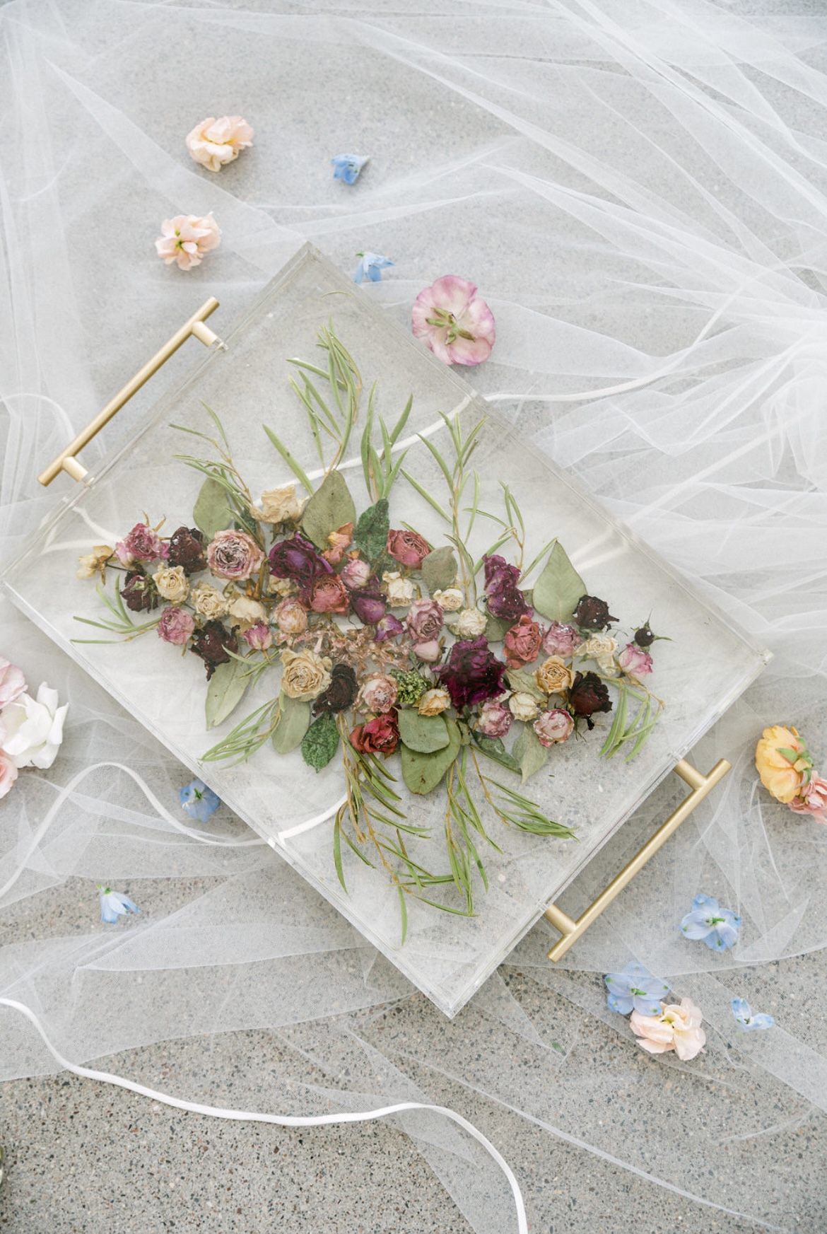 Flat arrangement of pink, purple, and green flowers on a white rectangular tray, with scattered small flowers and tulle fabric around.