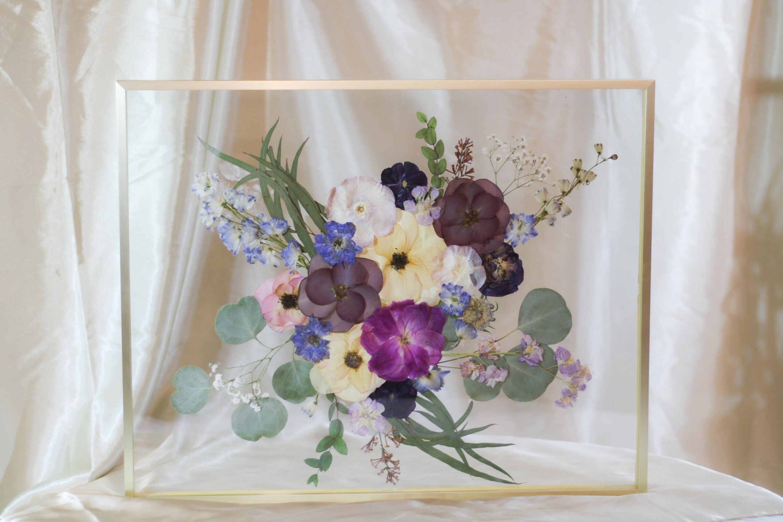 Flower arrangement with purple, white, and blue flowers in a glass box, against a light curtain background.