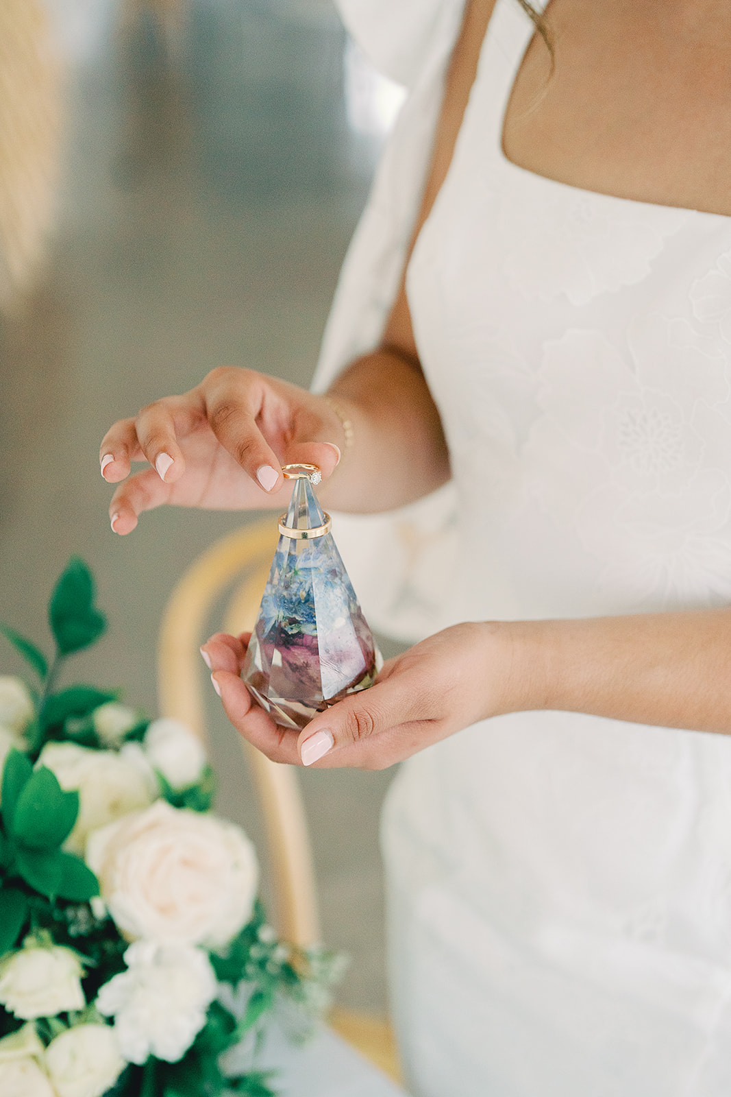 Person in white dress holding a glass perfume bottle with a floral arrangement in the background.