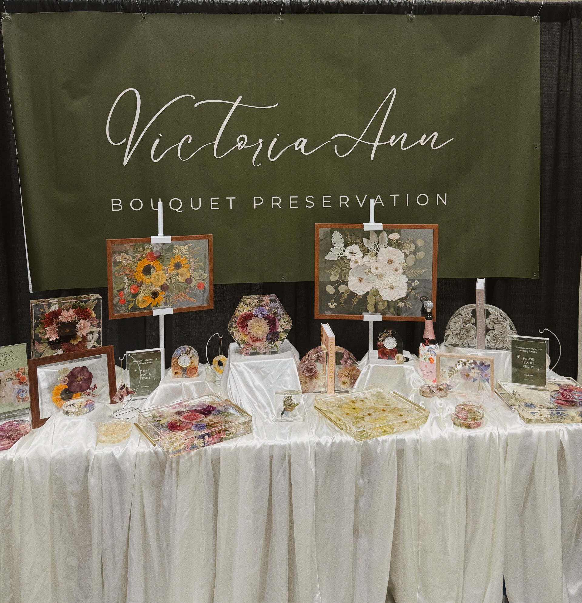 Display table with floral artwork and decorative items in front of a green banner reading Victoria Ann Bouquet Preservation.