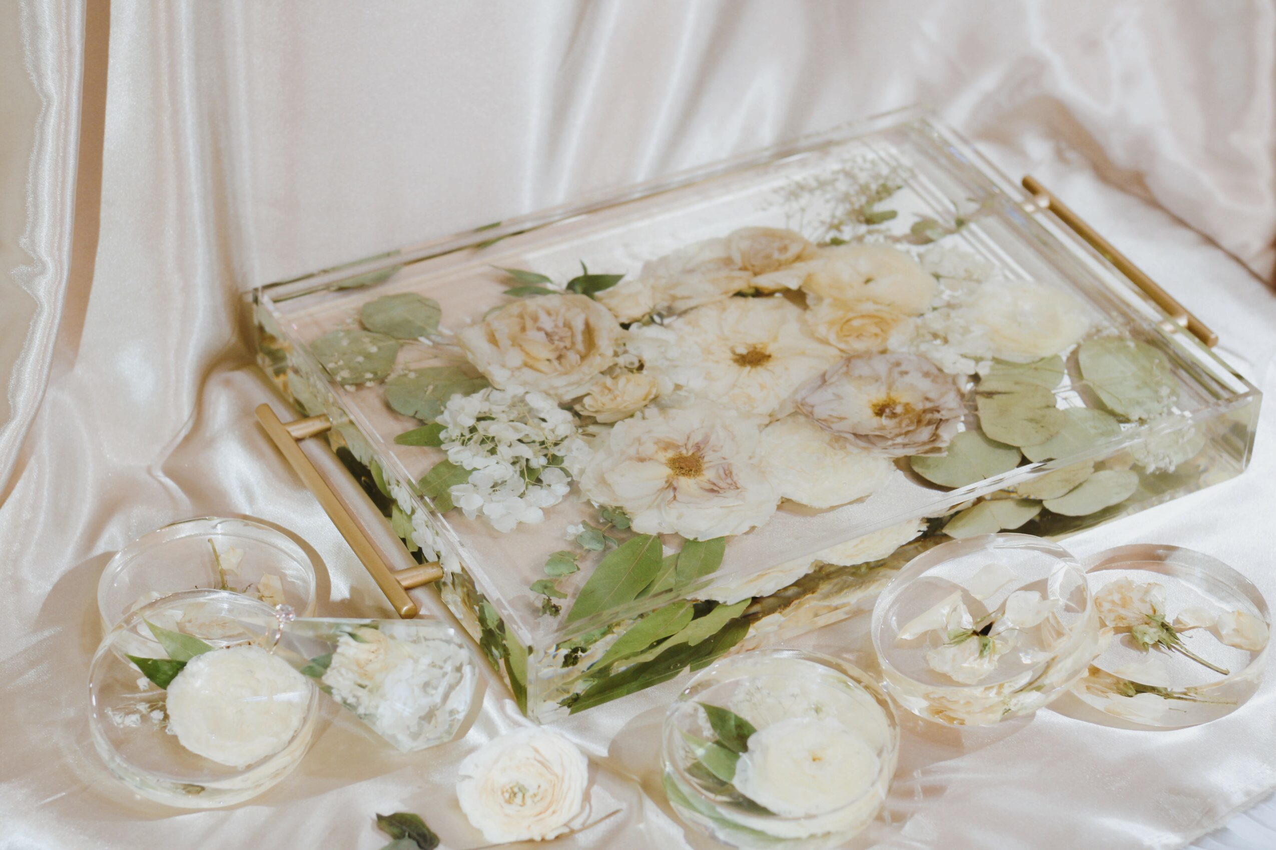 Decorative glass tray with white flowers and green leaves, surrounded by small glass bowls with white floral arrangements, on satin fabric