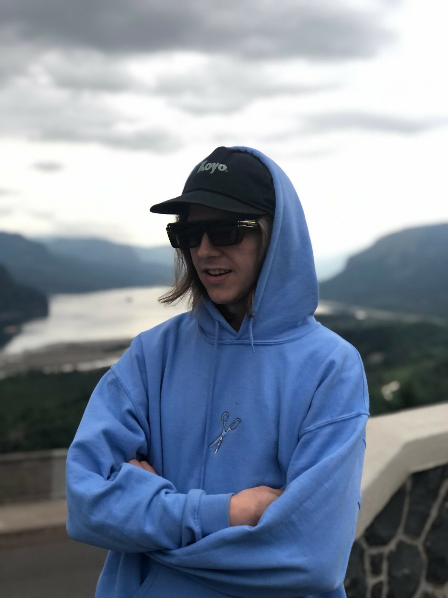 Person wearing a blue hoodie, sunglasses, and a cap outdoors with mountains and a body of water in the background.