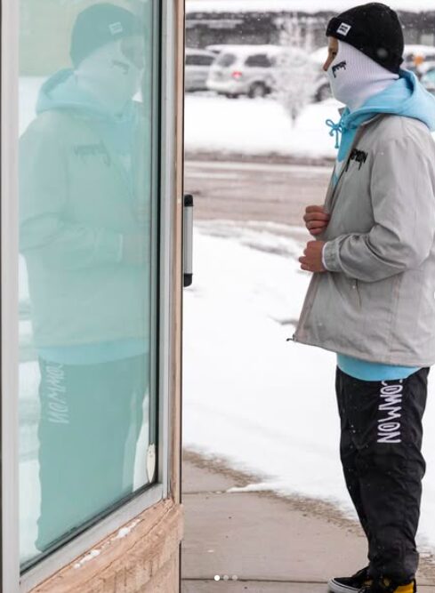 Person in winter clothing stands outside near a glass door, snow on the ground, and a reflection of the person in the glass.