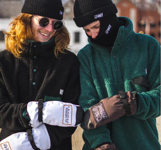 Two people wearing winter clothing, hats, and gloves, smiling and looking at each other outdoors.