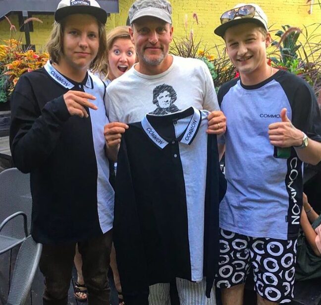 Four people, three young boys and an adult man, stand outdoors holding a black and white shirt. They smile at the camera.