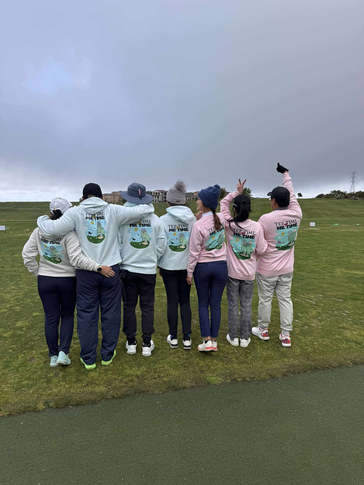 Group of eight people standing on grass, facing away, with cloudy sky above, some making peace signs, wearing matching hoodies.