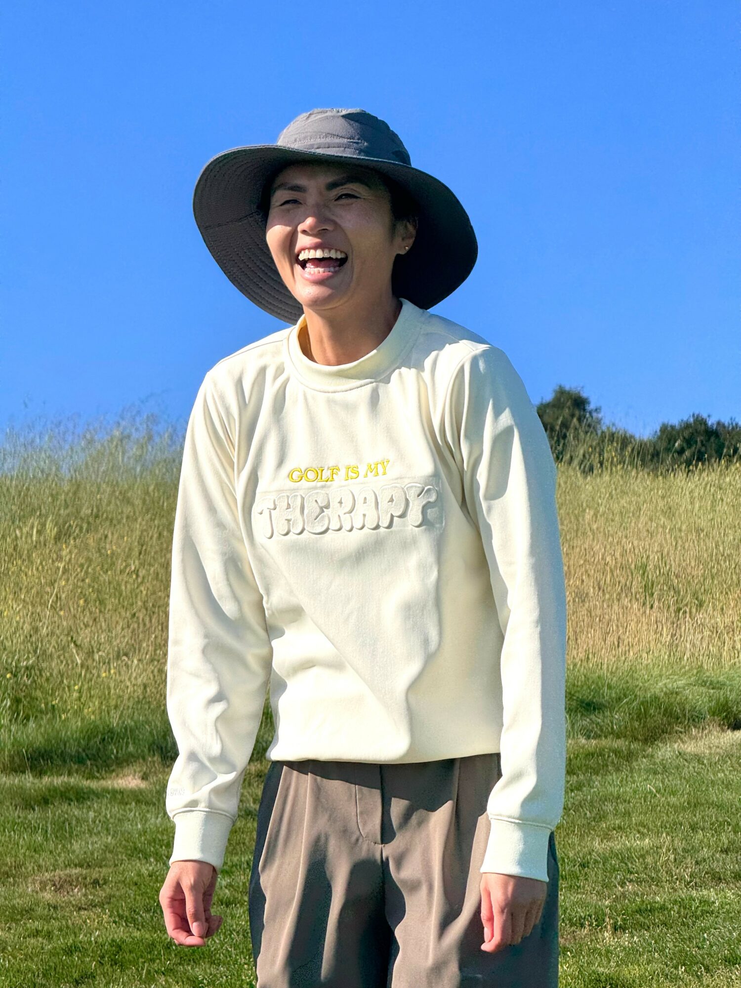 Woman smiling outdoors wearing a wide-brimmed hat and light-colored sweatshirt, standing in a grassy field under a blue sky.