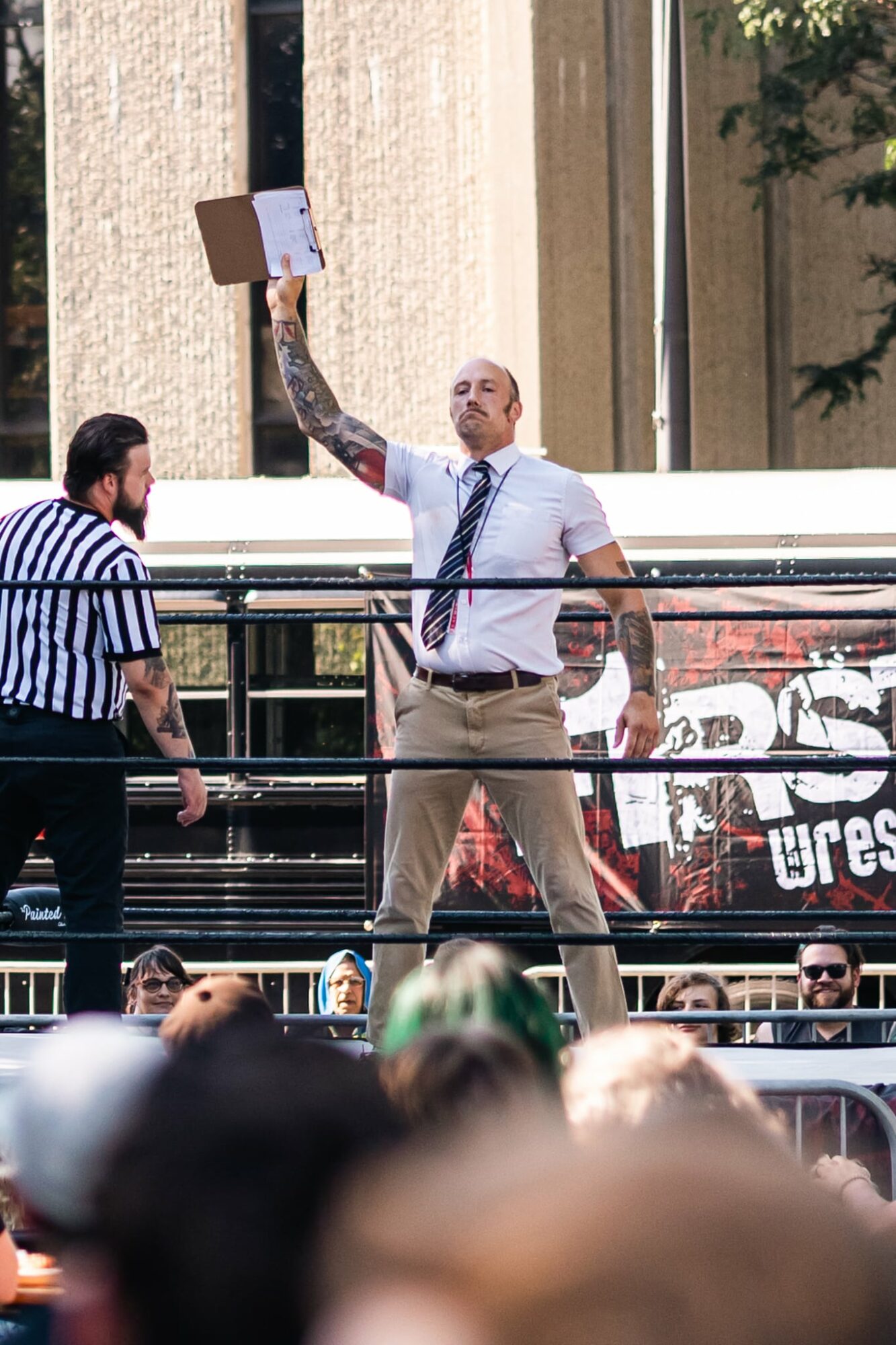 Man in white shirt and tie holding a cardboard sign in a wrestling ring, with referee and audience around.