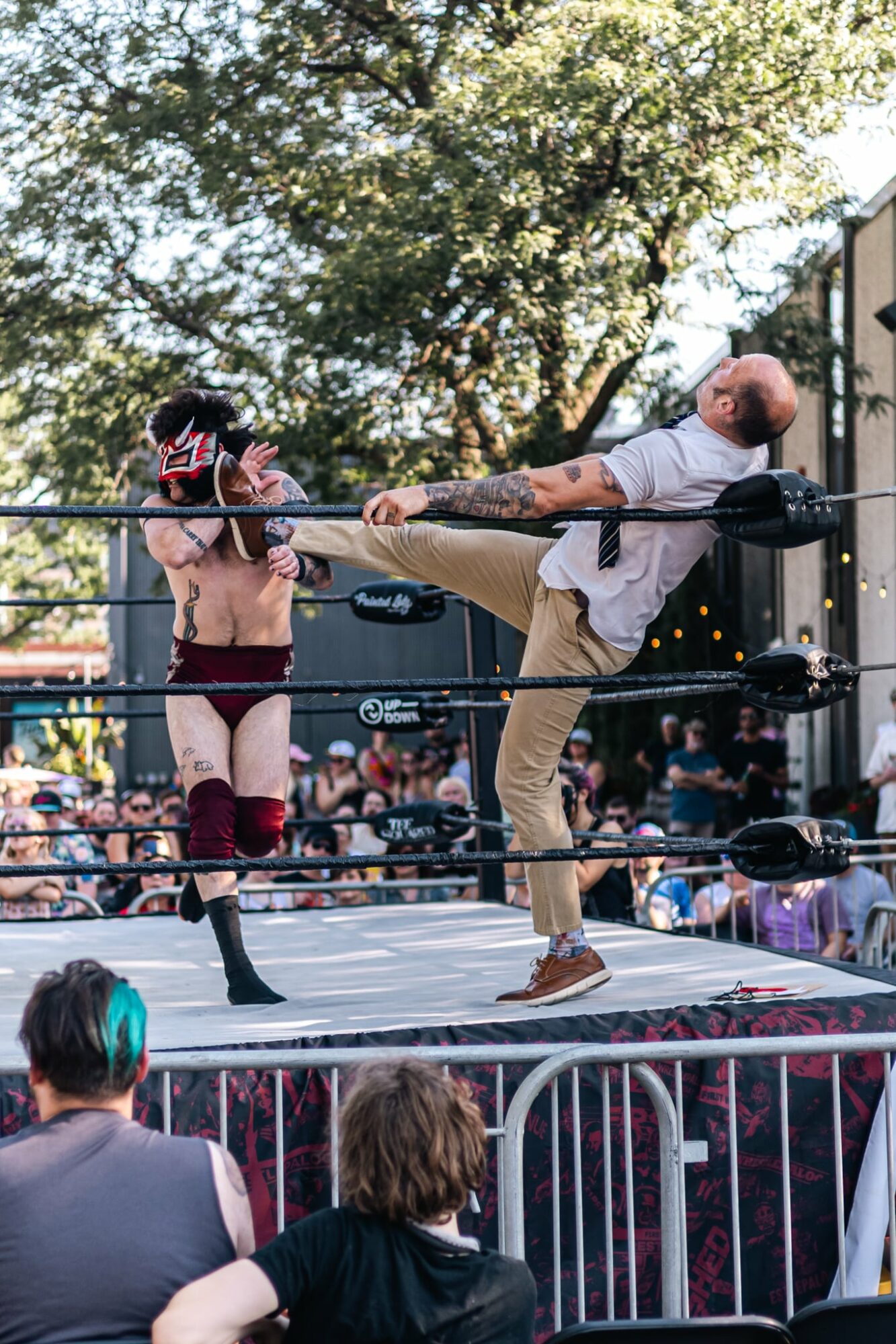 A wrestling match with one wrestler kicking another in the face inside a ring, audience watching outside, outdoors with trees.