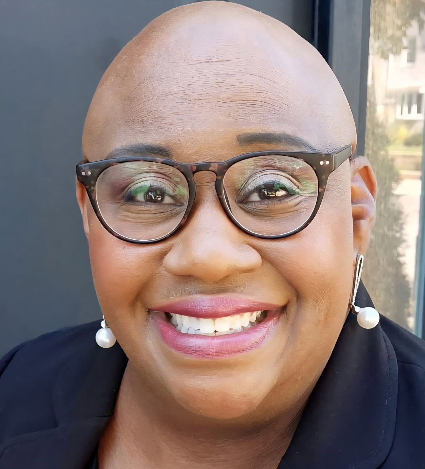 Close-up of a smiling person with glasses, earrings, and a bald head, indoors near a window.