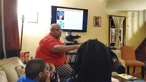 A man in a red shirt and glasses is sitting at a table, speaking to a group in a living room with a TV and window.